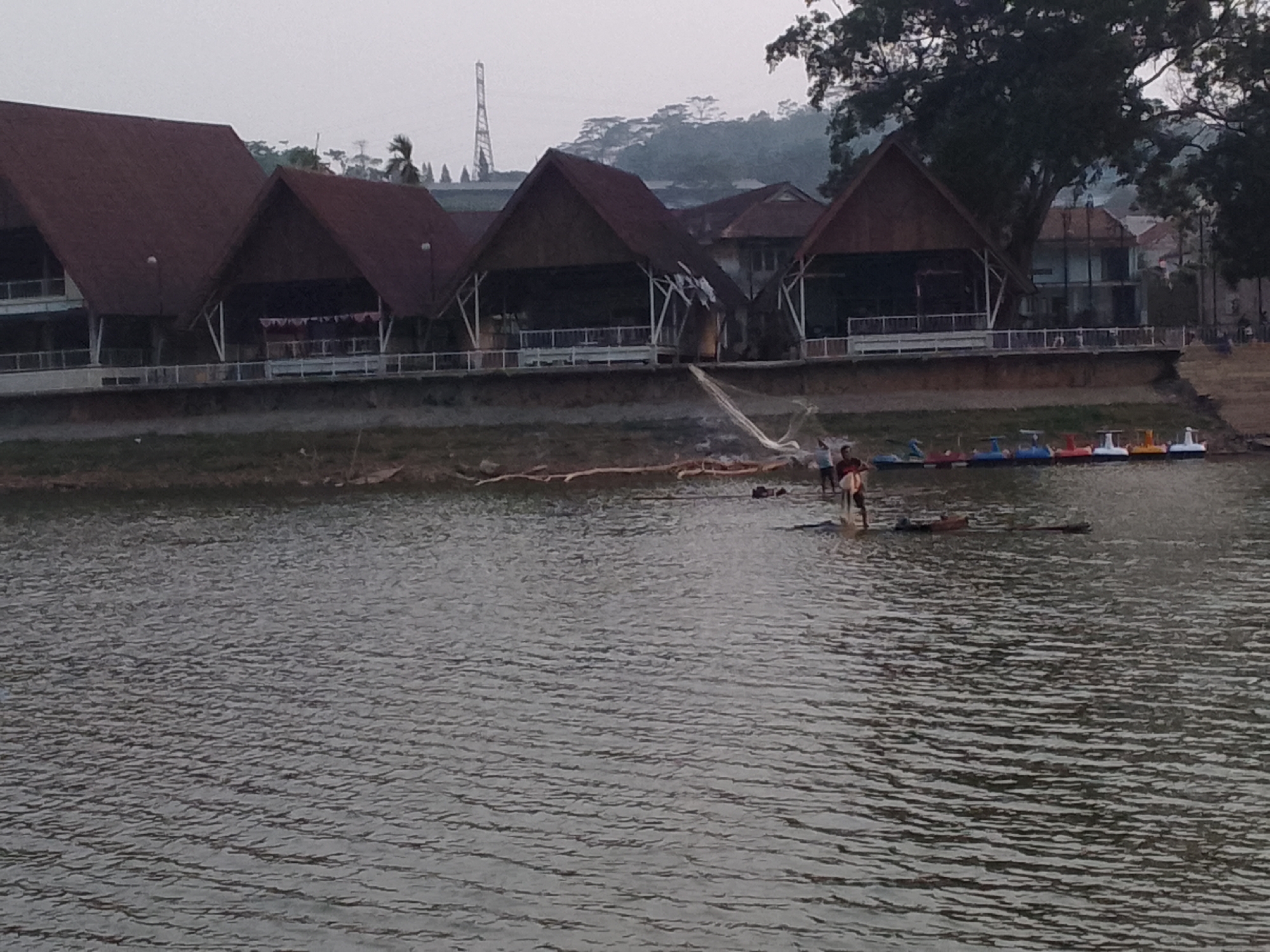 In the city of Ciburuy, Indonesia, a wide body of water stretches across the foreground, reflecting the overcast sky with textured ripples. Along the far bank, a row of brown-roofed buildings with open, pavilion-like structures are visible. These buildings are elevated on stilts and feature railings. In the water near the shore, several small, colorful paddle boats are moored.
A group of individuals are present in the water. Two people are standing near the bank, and one appears to be casting a fishing net. Further out in the water, another person is on a raft-like object. The environment suggests a recreational area or a lakeside park. The background shows a gentle slope of land covered in vegetation and trees, with some houses and a tall, lattice tower visible in the distance. The overall atmosphere is calm and somewhat hazy.