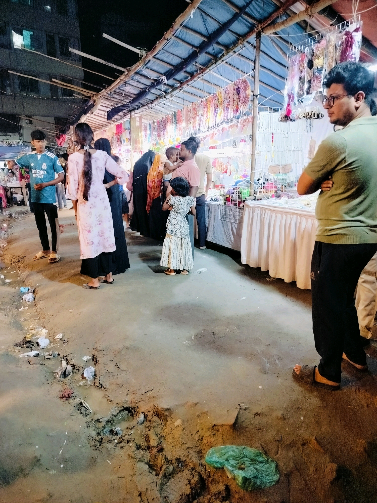 This image depicts an outdoor market scene at night, likely in Dhaka, Bangladesh, characterized by temporary stalls and a rough, muddy ground surface. A young boy wearing a blue t-shirt with "Emirates Fly Better" printed on it stands on the left. To his right, a woman in a pink floral-patterned outfit with a long braid is visible. Several other individuals are present, including a woman in a black abaya, a woman in an orange hijab holding a baby, and a small child in a patterned dress. They appear to be browsing or interacting at the market stalls, which are laden with various goods, including jewelry, accessories, and possibly toys. The stalls are covered with tarpaulin or similar material, supported by bamboo poles. The ground is uneven, with visible puddles of water and scattered debris, suggesting it may have recently rained or is a poorly maintained area. The lighting is artificial, emanating from the stalls and possibly nearby buildings, casting shadows and highlighting the textures of the environment. The overall impression is of a bustling but somewhat unkempt nighttime market.