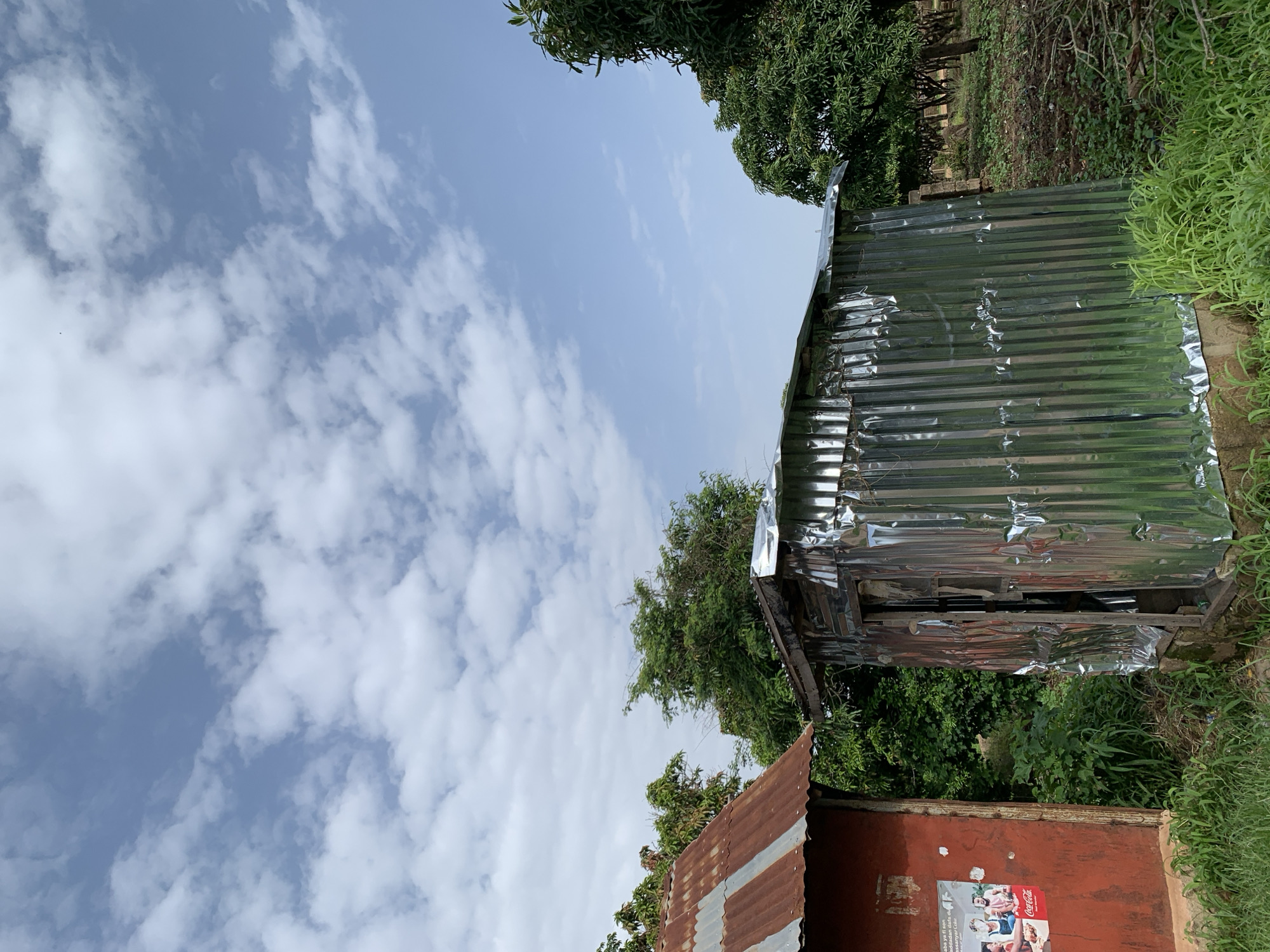 A photograph taken in Jos, Nigeria, shows two structures on the left, one with a rusty corrugated iron roof and a red plastered wall, and another adjacent structure with a corrugated aluminum exterior. The red-walled structure has a poster on its left side featuring a Coca-Cola advertisement with people. The aluminum-clad structure appears to be a small, makeshift building with an open doorway. Behind the structures, lush green trees and foliage are visible, suggesting a semi-rural or peri-urban environment. The sky above is a bright blue, dotted with scattered white clouds. The ground in front of the buildings is covered with grass and some scattered debris.