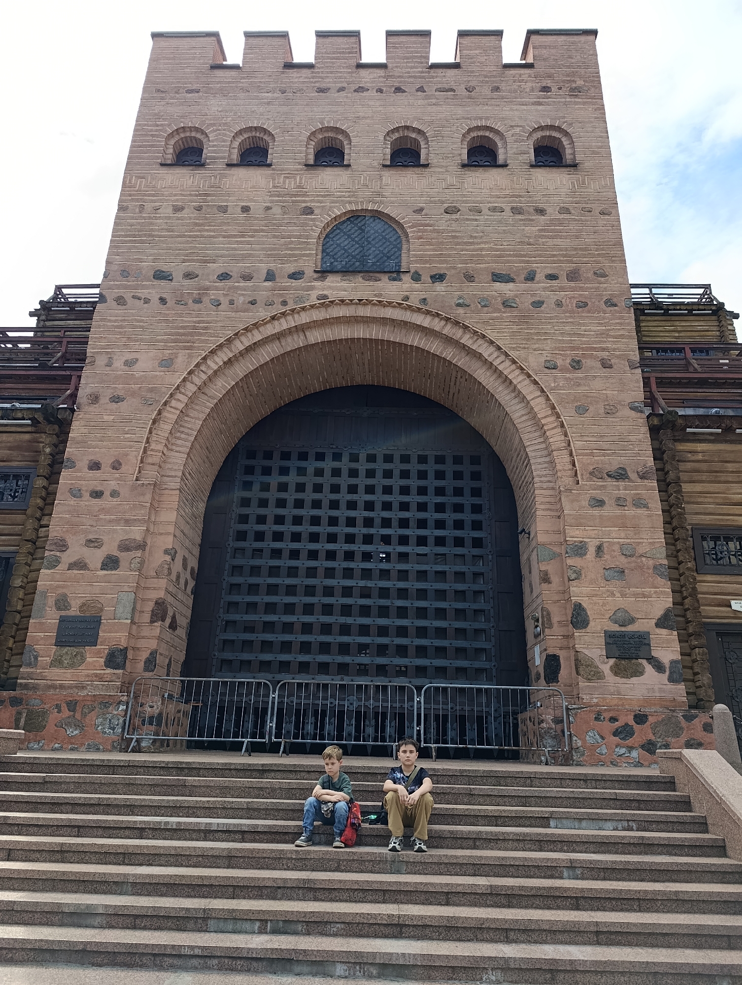 Two young boys sit on the wide granite steps of a large, imposing brick structure, possibly a historical building or fortress, in Kyiv, Ukraine. The building features a massive, dark metal gate with a grid pattern, flanked by stone and brickwork. Above the gate, arched windows are visible, and the structure is topped with crenellations. The steps themselves are a prominent feature, leading up to the entrance. The boys are casually dressed, one with a red backpack beside him, and the other with a green messenger bag. The overall scene suggests a tourist or recreational visit to a landmark. The sky above is partly cloudy, indicating daytime.