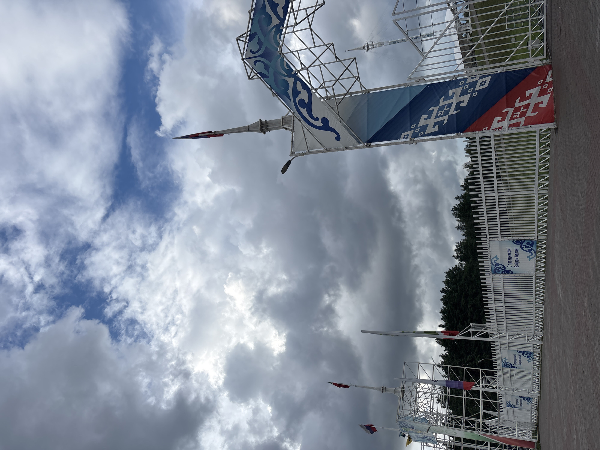 The image depicts the entrance gate to a festival or event in Naberezhnye Chelny, Russia, likely on an overcast day, given the dramatic cloud formations. The entrance is adorned with a large, arching metal structure decorated with blue and white swirling patterns, reminiscent of traditional Tatar motifs. A tall banner hangs from the right side of the arch, featuring diagonal stripes of red, blue, and white, with white geometric patterns.
To the left of the main entrance, a white metal fence stretches across the scene. Several flags are visible on tall poles behind the fence, including what appears to be the Russian flag and other regional flags. Some of these flags are partially obscured by the metal lattice structure of the entrance.
The fence itself has decorative blue and white banners attached at intervals, displaying text that reads "С Праздником! Байрам белән!" which translates to "Happy Holiday! With Holiday!" in Russian and Tatar, respectively. This confirms the festive nature of the location.
In the background, a dense line of evergreen trees forms a backdrop, suggesting the venue is located on the outskirts of the city or in a park-like setting. The ground in the foreground is paved with reddish-brown interlocking bricks. No people are visible in the image. The overall impression is of a welcoming and festive entrance to an outdoor celebration.