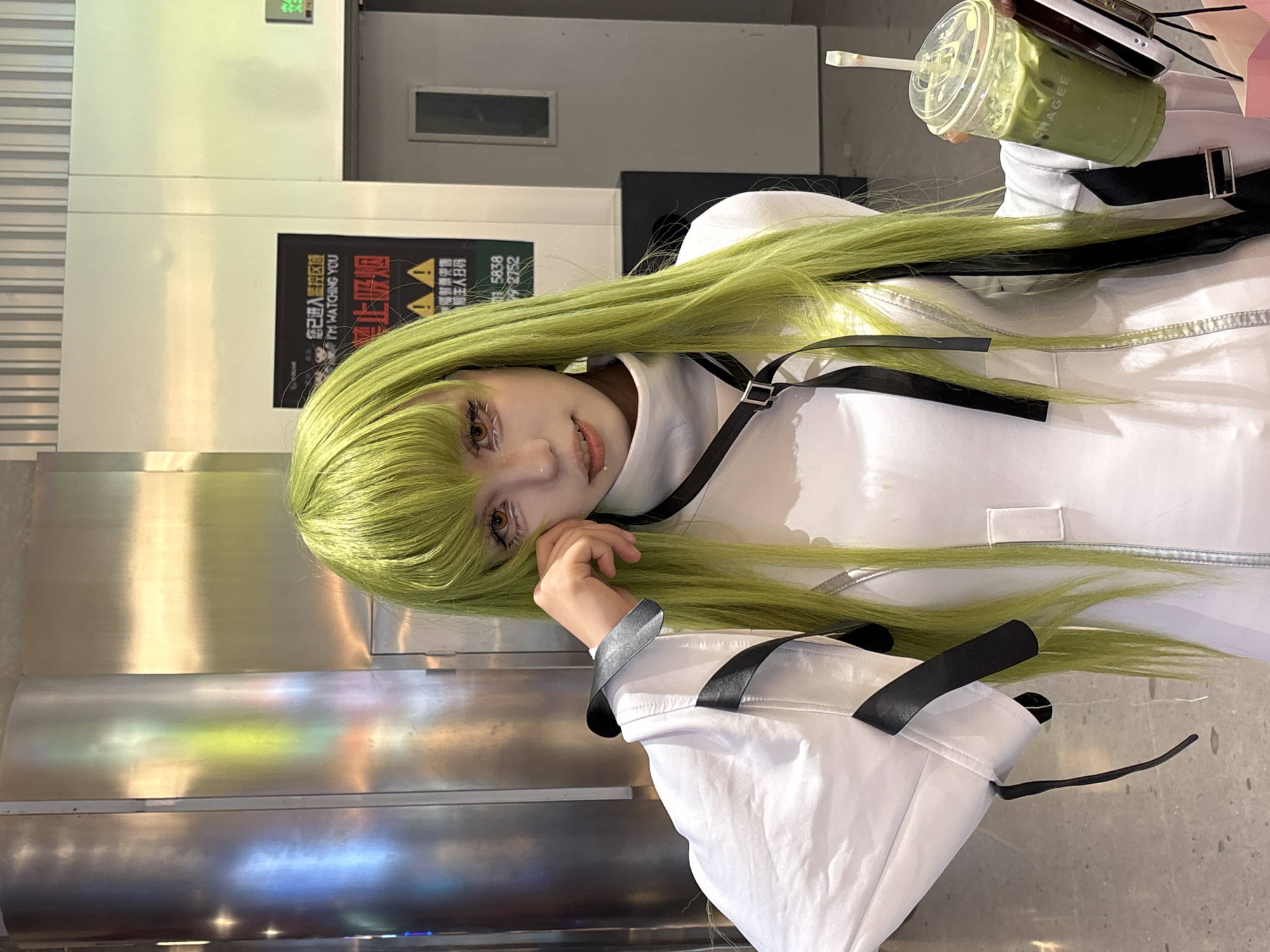 A person in a white and black outfit with long, light green hair is indoors, holding a drink from CHAGEE in the upper right corner. A sign on the wall includes the phrases "I'M WATCHING YOU" and "禁止吸烟," which translates to "no smoking," along with phone numbers "15838" and "2752." The person is in Liangjiang, China, as indicated by the Chinese text on the sign. The background includes elevator doors.