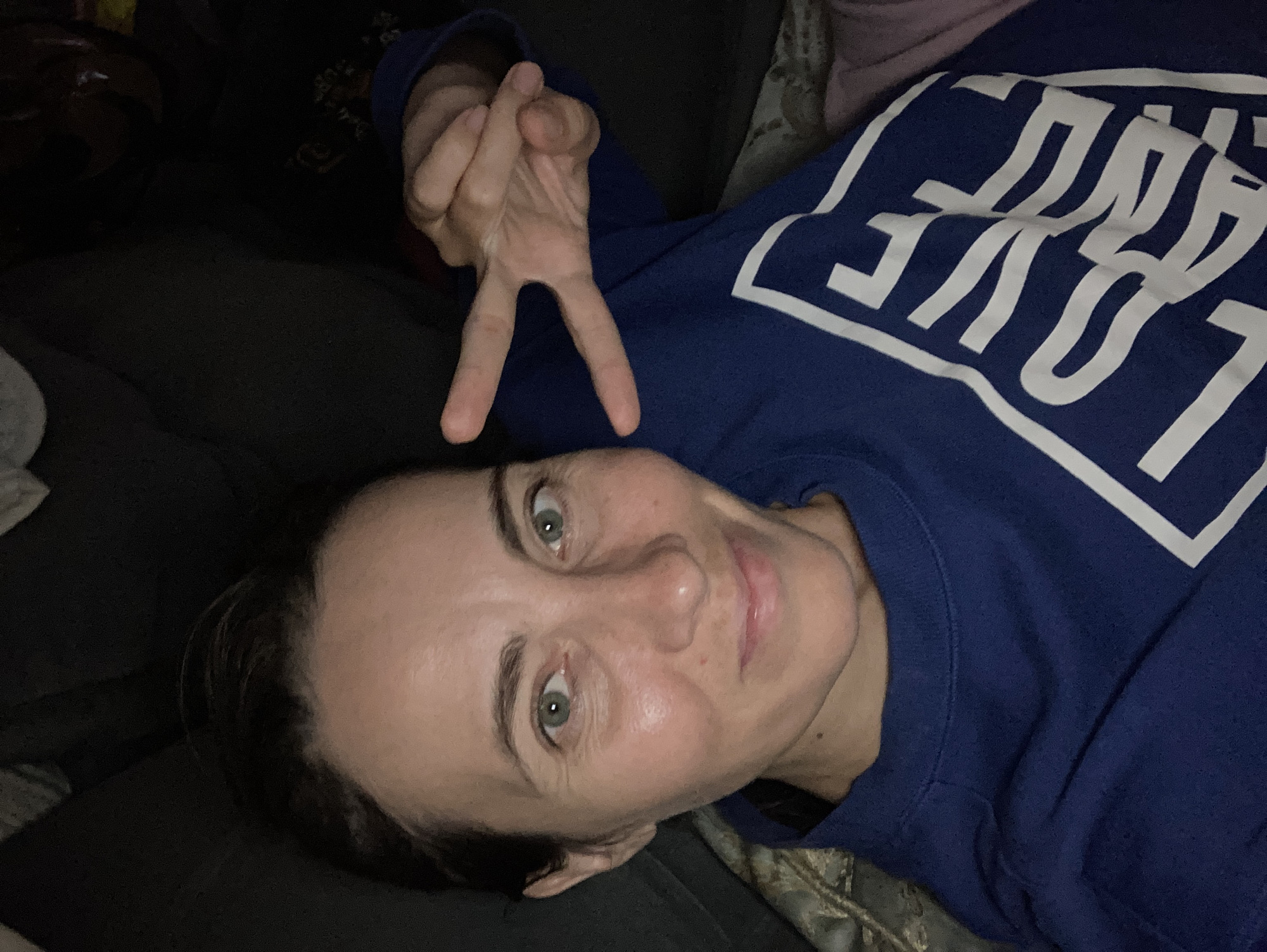 A woman lies indoors, looking directly at the camera with a serene expression, her head tilted slightly. She has fair skin, dark hair, and striking green eyes, which are wide open and engaged. A faint smile plays on her lips, suggesting a calm or content mood. Her right arm is raised, forming a peace sign (V-sign) with her index and middle fingers.
She is wearing a dark blue, long-sleeved sweatshirt featuring a prominent, stylized white text on the chest that clearly spells out "LOVE," enclosed within a bold white rectangular outline. The background is softly lit and consists of dark, textured fabrics, likely a couch or bedding, with a lighter, patterned fabric visible behind her head. The dim lighting suggests the photograph was taken during the evening or night. The overall scene conveys a relaxed, comfortable atmosphere within a private indoor setting, situated in Florianópolis, Brazil.