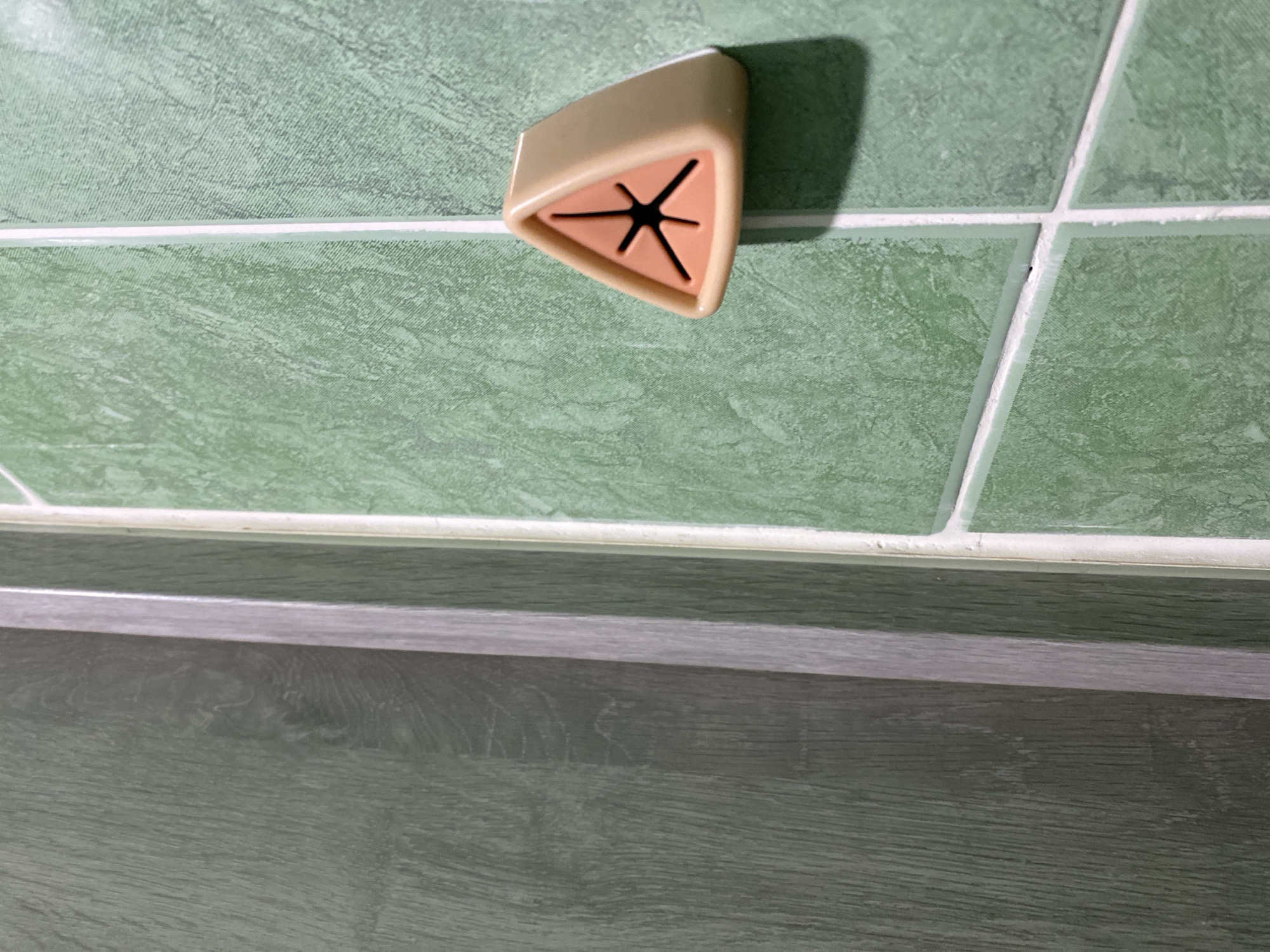 A triangle-shaped, beige towel holder with a dark red starburst pattern on its face is mounted on a green tiled wall with white grout. The wall appears to be above a wooden surface. The location is Ternopil, Ukraine.