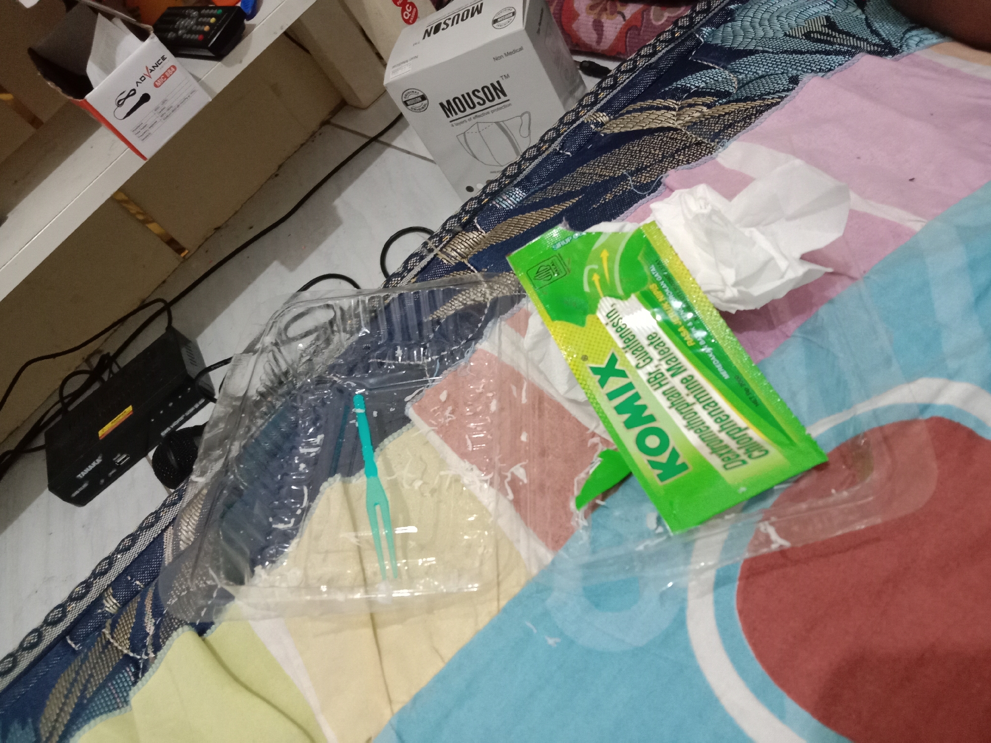 The image displays a domestic scene inside a room, likely a bedroom, with a focus on several objects laid out on a mattress. The overall lighting suggests a dimly lit indoor environment, possibly from a low-wattage bulb or ambient light. In the foreground, a green sachet labeled "KOMIX" with text in Indonesian is prominently featured, suggesting it contains medication, likely cough syrup. Next to the sachet is a crumpled white tissue. To the left, a transparent plastic container, possibly for food or a medical kit, holds a small, bright teal plastic fork. Behind these items, a black electronic device with the brand name "TAKARA" is visible, connected by cables. Further in the background, a white box with the brand name "MOUSON" is visible, which appears to be packaging for face masks, indicated by the text "4 layers of effective protection." The objects are situated on a patterned mattress with blue, white, and gold woven fabric. To the left of the mattress, a wooden shelf holds another small electronic device with buttons and a power cord. The flooring is not clearly visible but appears to be a light-colored tiled surface. The scene is static, with no individuals present or engaged in any activity. The location is identified as Brangsong, Indonesia, implying a residential setting within that geographical area.