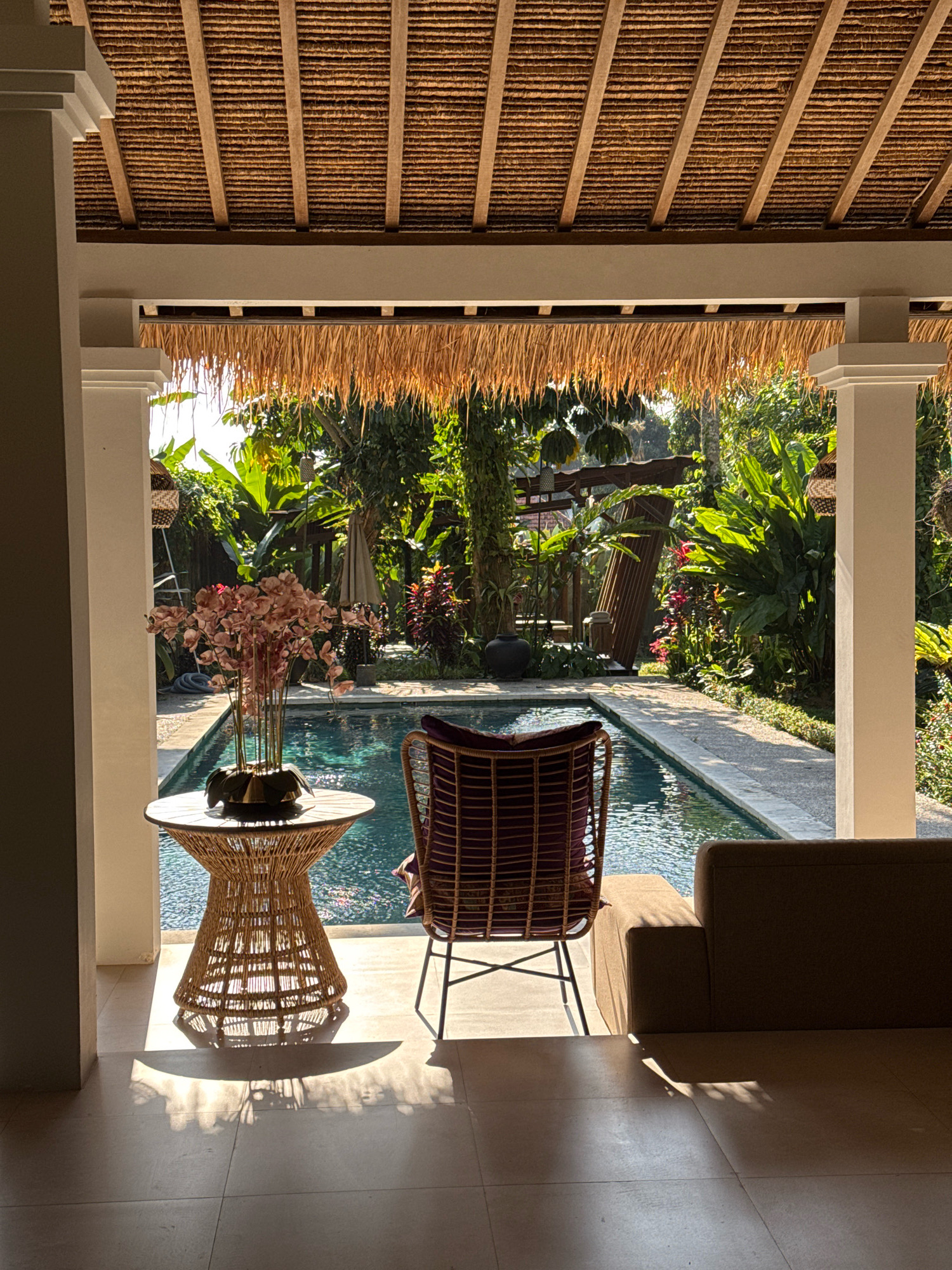 This image depicts a serene outdoor living space adjacent to a swimming pool, likely located in Tegallalang, Indonesia, characterized by its lush tropical vegetation and traditional thatched roofing. The scene is captured from a covered patio, looking out towards a bright, sun-drenched swimming pool.
In the foreground, to the left, a woven rattan side table holds a decorative vase filled with pinkish-red artificial flowers. The table casts a distinct shadow on the tiled floor, dappled with sunlight filtering through the overhead structure. To the right of the table, a single chair with a dark, woven frame and purple cushions sits facing the pool. Further to the right, a portion of a neutral-colored sofa is visible, also casting a shadow.
The central focus of the mid-ground is the inviting swimming pool, its surface reflecting the sunlight and the surrounding greenery. A narrow concrete path borders one side of the pool, leading into the garden.
The background is dominated by dense, vibrant tropical foliage, including large-leafed plants and trees, indicative of a warm climate. The roof structure above the patio is constructed from natural materials, with visible wooden beams and a thatched covering, topped with a fringe of dried palm leaves hanging down. The overall ambiance is one of tranquil relaxation, bathed in natural light. No people are visible in the frame.