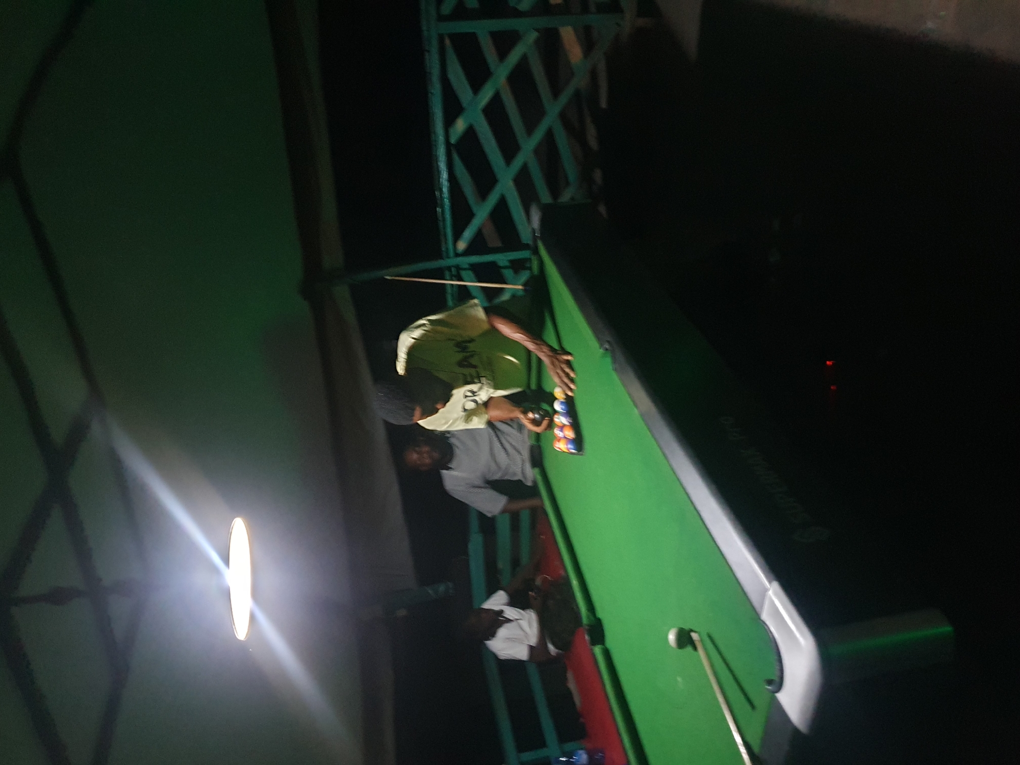 The image depicts a group of people gathered around a pool table in what appears to be an outdoor or semi-outdoor recreational area, possibly at the Mount Patti hotel in Lokoja, Nigeria. In the foreground, a person wearing a yellow t-shirt with the word "DREAM" is in the process of playing pool, with their hand positioned over the cue ball and a rack of pool balls visible on the table. The pool table itself is green and shows the branding "SUPERMAX Pro." To the left, another person in a white shirt is seated, observing the game, and behind them, a third person in a striped shirt is also watching. The environment is dimly lit by a ceiling light fixture, and a green railing is visible in the background, suggesting an elevated or enclosed space. The overall impression is a casual gathering for a game of pool.