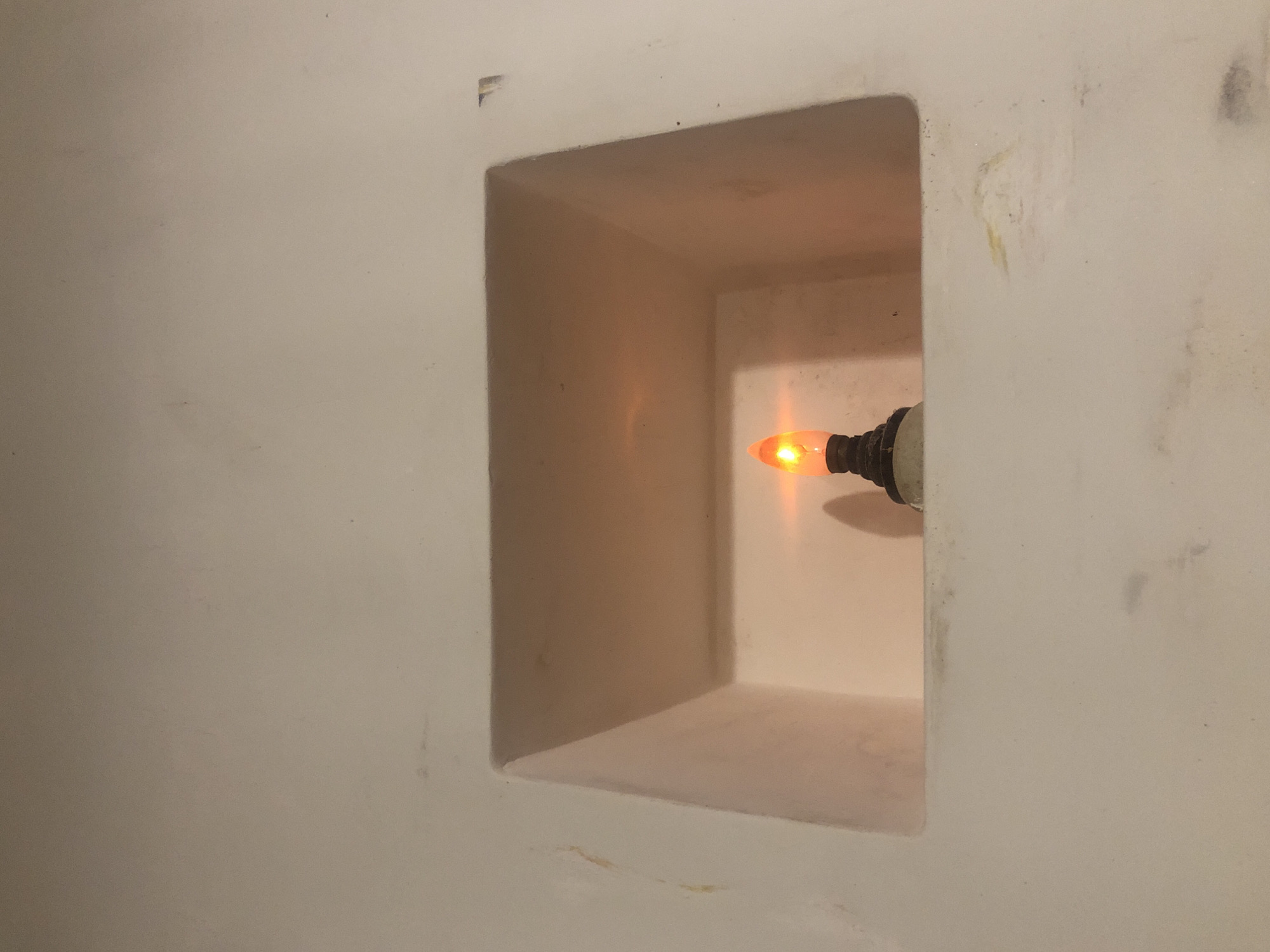 The image presents a close-up view of a small, orange-glowing light bulb situated within a recessed rectangular niche in a light-colored wall. The bulb emits a warm, soft, orange light and is screwed into a simple, light-toned fixture, positioned towards the right side of the niche.
The wall and the interior of the niche are painted or plastered in an off-white or very light beige color. The broader wall surface surrounding the niche shows several minor imperfections, including faint yellowish streaks, smudges, and darker marks, particularly visible on the right and bottom-left areas. A small chip or rough patch is discernible on the top-left outer edge of the recessed opening. The interior surfaces of the niche appear somewhat cleaner and are warmly illuminated by the bulb's glow.
No people are visible, and no specific activity is taking place other than the light bulb being illuminated. The glow of the bulb suggests the image was taken during a time of low ambient light, such as evening or night, or that the light serves as accent lighting. No discernible text is present in the image. Given the provided context, the scene is located in Ahangama, Sri Lanka.