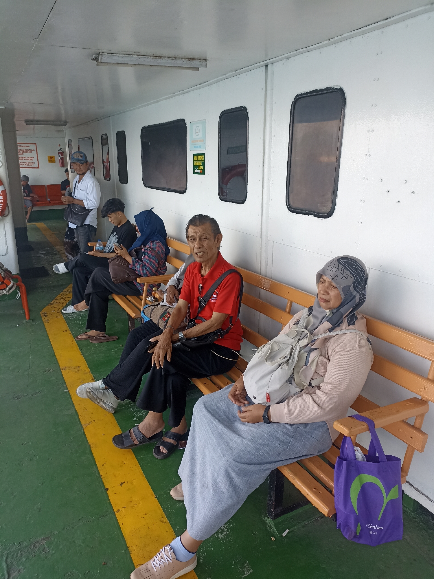 The image depicts a group of people seated on an orange wooden bench inside a ferry. The ferry appears to be docked or moving slowly, as indicated by the consistent lighting and the general atmosphere of waiting. The interior of the ferry is painted white with distinctive oval windows. A yellow and green striped line runs along the floor.
On the bench, from left to right, there are several individuals. The first visible person is an elderly man wearing a red polo shirt and black pants. He is looking towards the camera with a slightly open mouth. Next to him sits a woman wearing a grey hijab and a light pink long-sleeved shirt, with a light-colored backpack slung over her shoulder. She is looking to her left. To her left, a younger woman in a blue hijab and a plaid shirt is looking down at her phone. Beside her, a young man in a black t-shirt and black pants is also looking at his phone. Further to the left, a man in a white long-sleeved shirt and jeans stands, holding a black bag and looking to his left. In the background, other individuals can be seen sitting or standing, suggesting a moderate passenger load.
Notable objects include a purple tote bag with a green logo and text that reads "Chatime" hanging on the armrest of the bench to the right. Several water bottles are also visible on the bench. A life preserver is mounted on the wall to the far left. There are signs on the wall, one of which appears to be a "No Smoking" sign with a cigarette icon. The environment is utilitarian and functional, typical of ferry transport.
The scene is consistent with a ferry terminal or the interior of a ferry operating in Ketapang, Indonesia, which is known for its ferry services connecting to various islands. The attire of the individuals and the general ambiance suggest a local passenger group.
