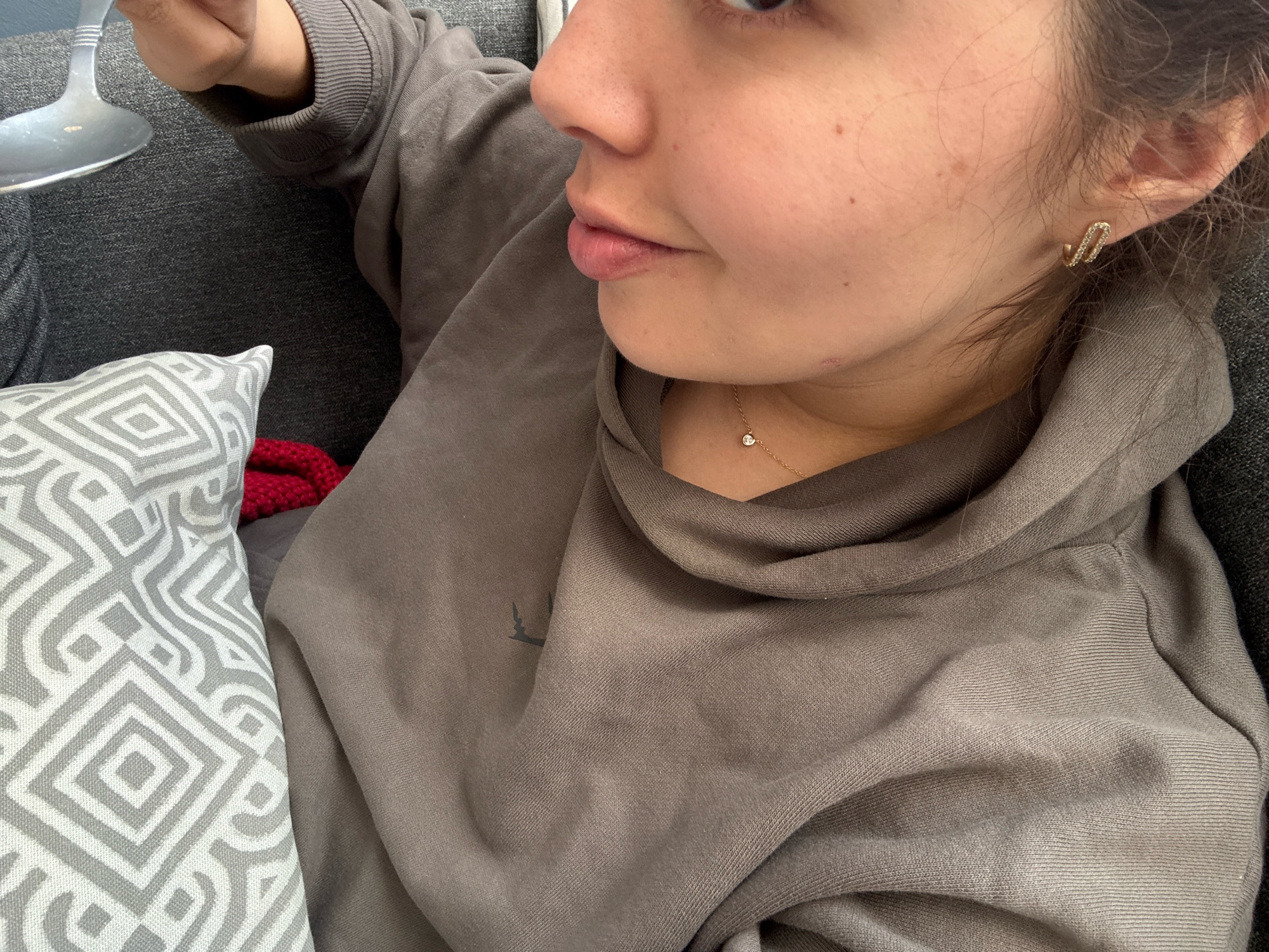 The image shows a close-up of a person's face and upper body, with a focus on their mouth, nose, ear, and shoulder. They are wearing a brown hooded sweatshirt and a delicate gold necklace with a small diamond pendant. A geometric-patterned pillow is visible in the lower left corner, partially obscuring a red textile. The person is holding a silver object, possibly a wine glass or a spoon, near their face. The background is a textured gray surface, likely upholstery. The scene appears to be indoors, possibly in a living room. There is no visible indication of the city of Puebla, Mexico.