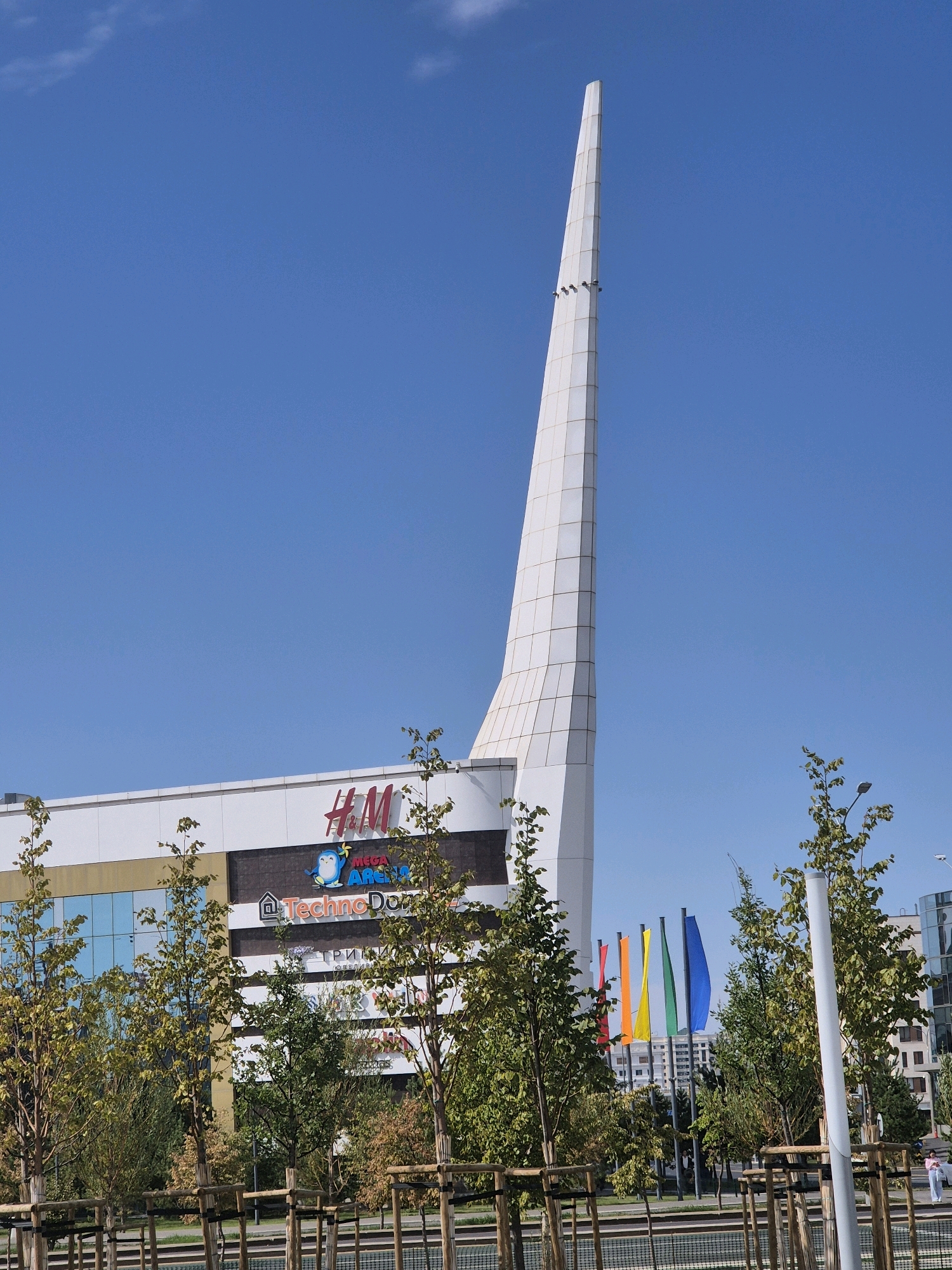 The image depicts a modern architectural structure with a tall, white, spire-like element, situated in the city of Astana, Kazakhstan. The spire is a prominent feature, rising against a clear blue sky with a few scattered white clouds. Below the spire, a building with large windows and a white facade is visible, featuring signage for "H&M" and "Mega Arena," as well as "TechnoDom." A row of young trees with light brown trunks and green foliage stands in front of the building, partially obscuring it. These trees are supported by wooden stakes. In the mid-ground, a set of colorful flags – red, orange, yellow, green, and blue – are displayed on poles. Further in the background, several modern buildings are visible, indicative of an urban environment. The overall scene suggests a commercial or entertainment complex in a well-developed area of Astana.