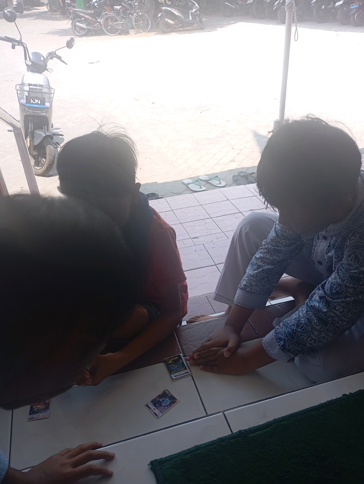 In Borobudur, Indonesia, two children are shown playing a card game. The scene appears to be an outdoor setting, possibly a patio or walkway, with a tiled floor and a sunlit background. Parked motorcycles are visible in the distance, suggesting a public area or a residential compound. One child, wearing a blue and white patterned shirt, is seated and actively engaged in the game, with their hands placed on the cards. The other child, in a red shirt, is also seated and appears to be playing. The image captures a moment of shared activity between the two children.