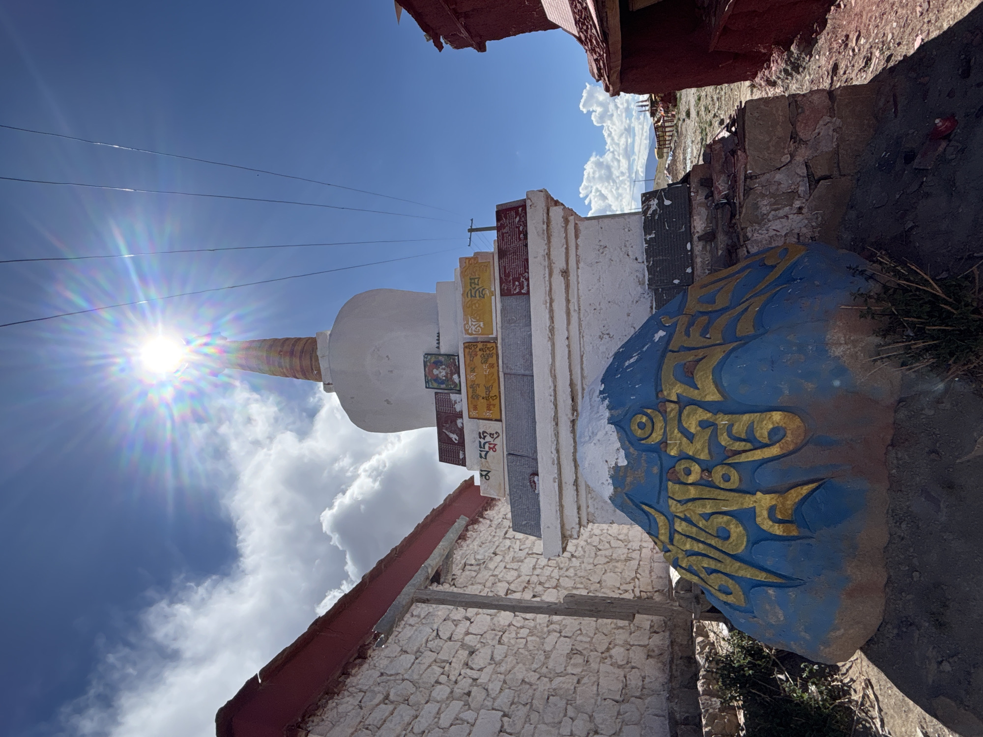 This vibrant outdoor image, captured in Labrang township, China, depicts a traditional white stupa (chorten) under a brilliant blue sky. The stupa, adorned with a golden spire, features a base decorated with numerous rectangular panels. These panels display a mix of sacred images, possibly deities or mandalas, and intricate Tibetan script in various colors, including clear black text on a yellow panel and a red panel.
Positioned prominently in front of the stupa is a large, irregularly shaped rock, boldly painted blue with striking golden Tibetan characters, likely a religious mantra. The stupa is bordered on either side by rustic structures, characterized by whitewashed stone walls and traditional reddish roofs or wooden overhangs, suggesting an architectural style common in the region.
The time of day appears to be mid-day, with the bright sun positioned high in the sky, creating a pronounced sunburst lens flare in the upper left portion of the frame. The weather is sunny with scattered white clouds. Several thin power lines traverse the upper sky. Sparse vegetation is visible on the uneven ground near the painted rock. The scene conveys a sense of spiritual tranquility and cultural richness, characteristic of Tibetan-influenced areas.
