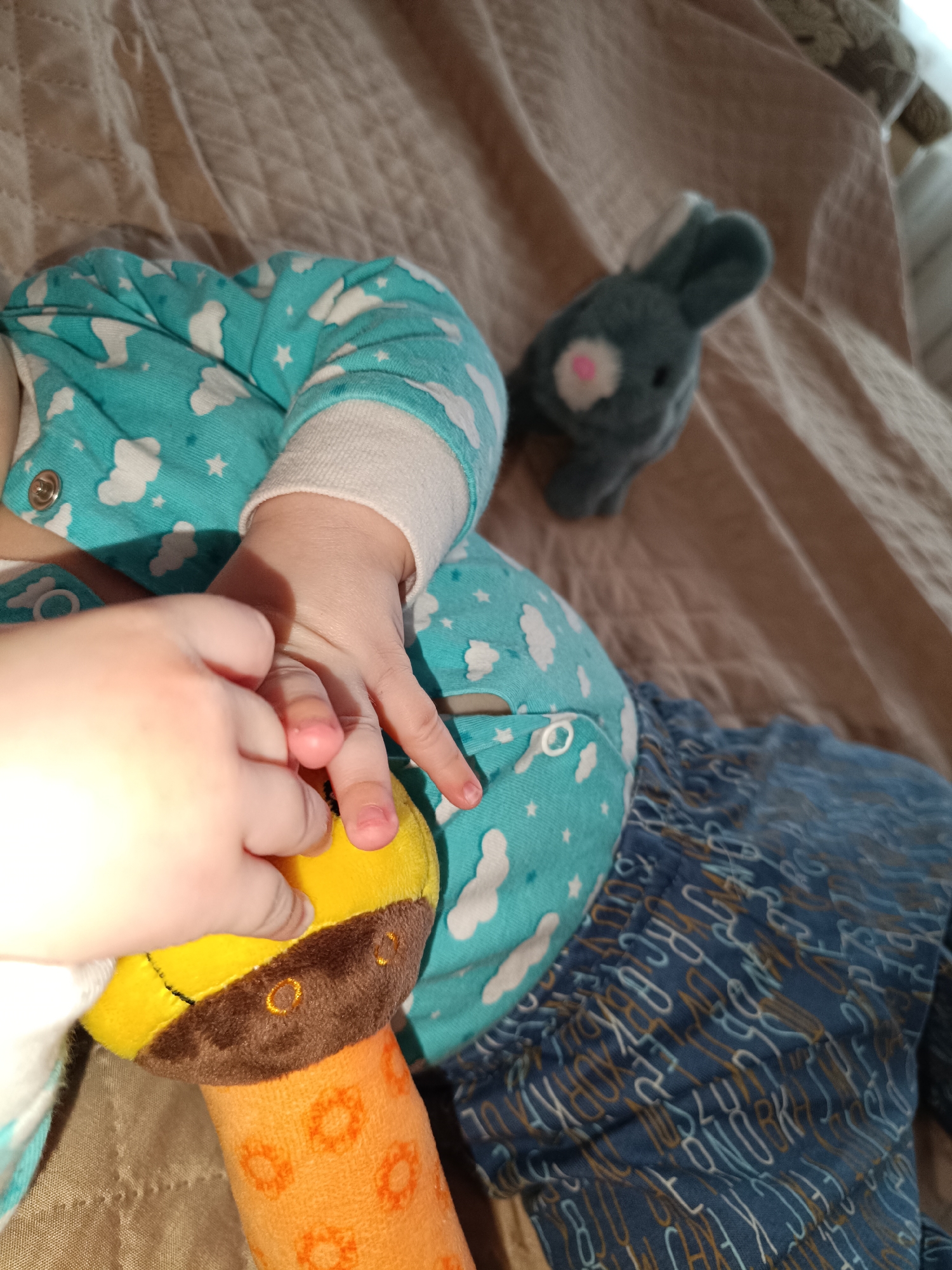 A baby is lying on a tan quilted surface, possibly a bed. The baby is wearing a turquoise pajama set with white clouds and stars. The baby is holding a yellow and orange stuffed giraffe toy with brown markings. A gray stuffed rabbit toy is nearby. The pajama bottoms are blue with a pattern of letters. The photograph was taken indoors, under natural lighting. There is no indication that the photo was taken in Tobolsk, Russia.