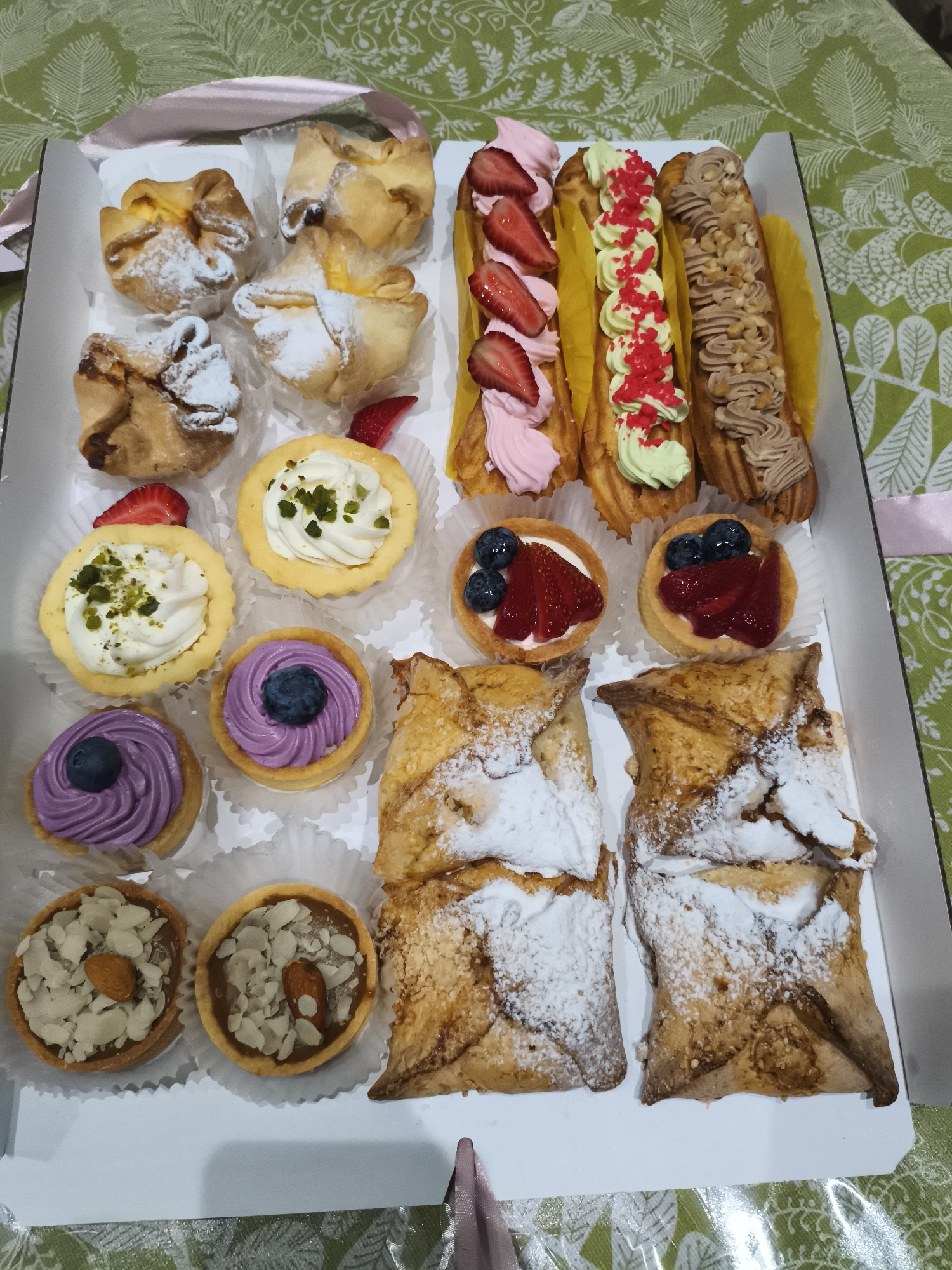 The image shows a white rectangular box filled with an assortment of pastries. The box is placed on a green tablecloth with a leaf pattern.
The pastries include several types:
* Four small, golden-brown baked goods, each folded into a square shape and dusted with powdered sugar.
* Three long, thin pastries resembling eclairs, filled with different colored creams and toppings. One is topped with pink cream and sliced strawberries. Another has light green cream and red sprinkles. The third is filled with brown cream and chopped nuts.
* Two small circular tarts with a creamy white filling and green specks, possibly pistachios.
* Two small circular tarts with a creamy white filling and topped with blueberries and sliced strawberries.
* Two small circular tarts with a purple swirled filling and a single blueberry on top.
* Two small circular tarts filled with a brown caramel-like substance and topped with sliced almonds and whole almonds.
* Two larger, square-shaped pastries dusted with powdered sugar, with a flaky, golden-brown exterior.
There are also a few sliced strawberries visible amongst the pastries. A light pink ribbon is visible tied around the top edge of the box, suggesting it might be a gift or part of a celebration. The overall scene is well-lit, suggesting it was taken indoors during the daytime. There is no visible text in the image. The arrangement of the pastries indicates they are likely for consumption or as a dessert offering.