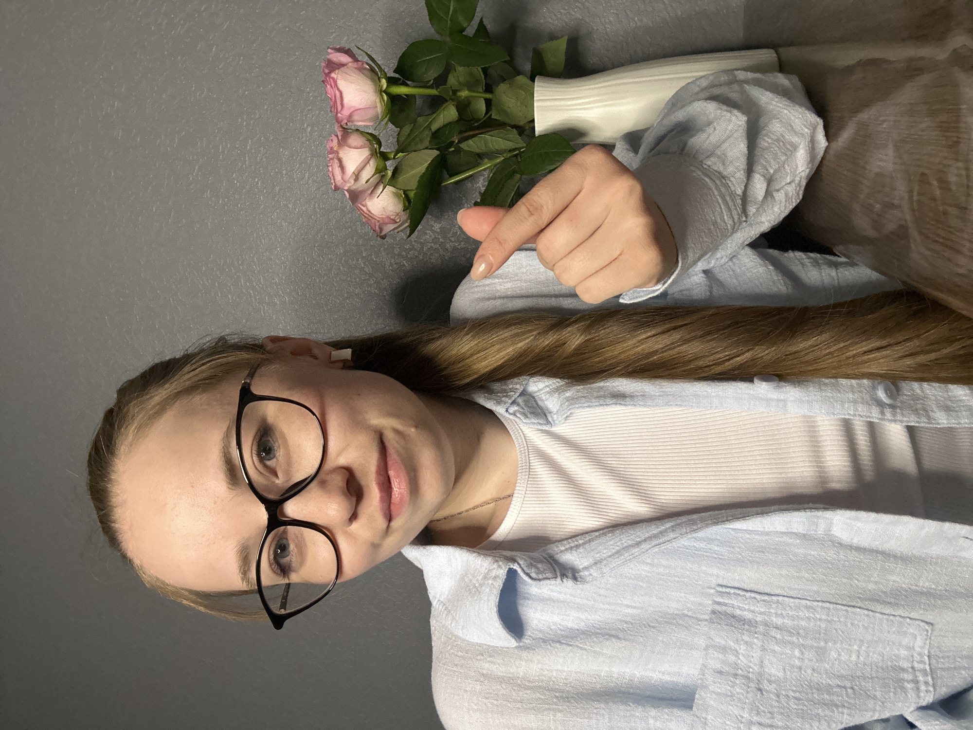 A young woman with blonde hair and wearing glasses is oriented toward the viewer. She has on a white ribbed tank top and a light blue collared shirt. Her right hand is extended toward a white vase with three pink roses. The background is a gray wall and a section of a wooden surface. There is no indication that this photo was taken in the city of Saint Petersburg, Russia.