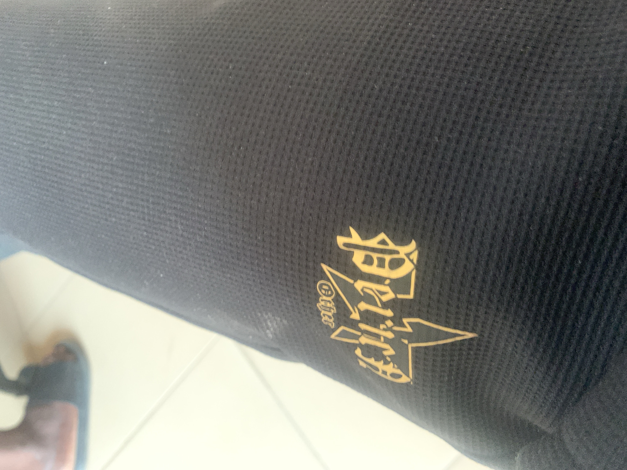 The image shows a close-up of a black garment with a yellow logo. The logo is stylized and appears to contain lightning bolt imagery along with lettering. The fabric of the garment has a textured, waffle-like pattern. In the upper left corner, out of focus, a person's foot wearing a dark-colored sandal is visible, positioned on a light-colored tiled floor. The overall scene suggests an indoor or partially shaded outdoor environment. There are no discernible elements within the image that specifically indicate the location as Jalingo, Nigeria.