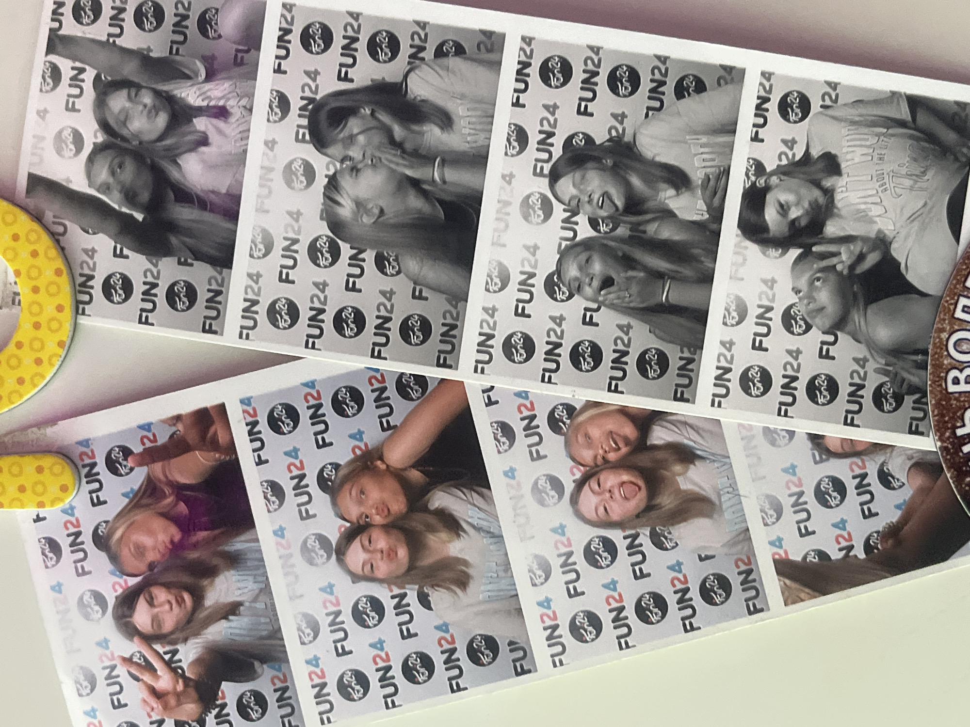 The image displays multiple photo booth strips arranged on a light-colored surface, capturing lively moments of young women. The strips feature various poses and expressions, some in black and white, and others in color, suggesting different photo booth sessions or options.
The primary subjects are several young women or girls, appearing to be friends, engaging in playful activities characteristic of a photo booth. They are seen making funny faces, pouting for kisses, sticking out their tongues, making peace signs, and laughing, all conveying a sense of happiness, joy, and camaraderie.
A consistent detail across all photo frames is the patterned background featuring the text "FUN24" and a corresponding circular logo with "Fun24" and a stylized '24' repeatedly printed. This branding points to a specific entertainment venue, located in Saint Petersburg, Russia, as per the provided context. On one of the color photo strips, an individual wears a light-colored t-shirt with text that reads "ABOUT THE UTA". Partially visible elements include a yellow, polka-dotted object on the left side of the main image and a brown object with the letters "BO" on the right, likely props or nearby decorations. The overall scene reflects a fun social event or gathering.