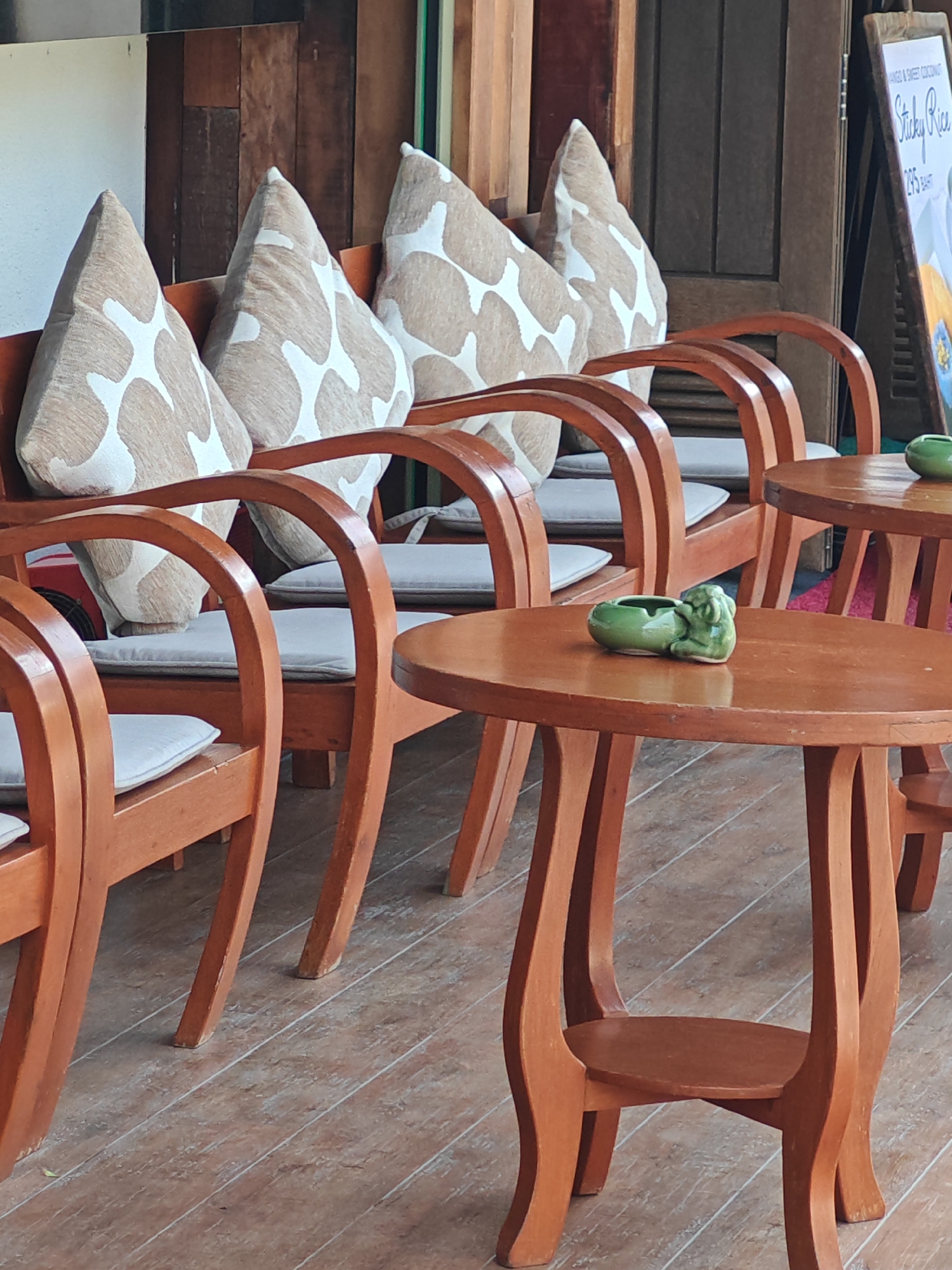 This is a medium-shot, eye-level, outdoor photograph of an empty seating area in Baan Bo Phut, Thailand. The scene features several wooden chairs with light-colored cushions, arranged around circular wooden tables. Decorative pillows with a tan and white pattern are placed on the chairs. On one of the tables, a small, green ceramic ashtray shaped like a frog is visible. The background consists of wooden walls and a partially visible menu board. The floor is made of wooden planks. No people are present in the image.