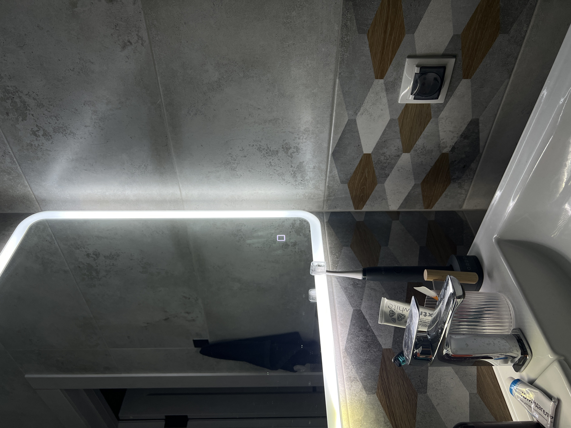 The image shows a close-up view of a modern bathroom sink area, likely located in Dudareva, Russia. The scene is dominated by a large, illuminated mirror with a rounded top edge. The wall behind the sink features grey tiles with a textured, concrete-like appearance, interspersed with decorative tiles in a geometric pattern of grey, white, and wood tones.
In the sink basin, a chrome faucet with a fluted base is prominent. Next to the faucet, in a clear fluted holder, are a toothbrush and a tube of toothpaste labeled "extra WHITE." To the right of this, a black electric toothbrush stands upright in its charger base.
On the left side of the sink, a tube of toothpaste is partially visible, with branding that appears to be "Parodontax." A black towel hangs on a hook reflected in the mirror on the left side of the frame.
On the right side of the wall, above the decorative tiling, a white electrical outlet with a dark cover is visible. The lighting in the bathroom appears to be artificial, primarily from the illuminated mirror, casting a bright glow. The overall aesthetic is contemporary and clean. There are no people visible in the image.