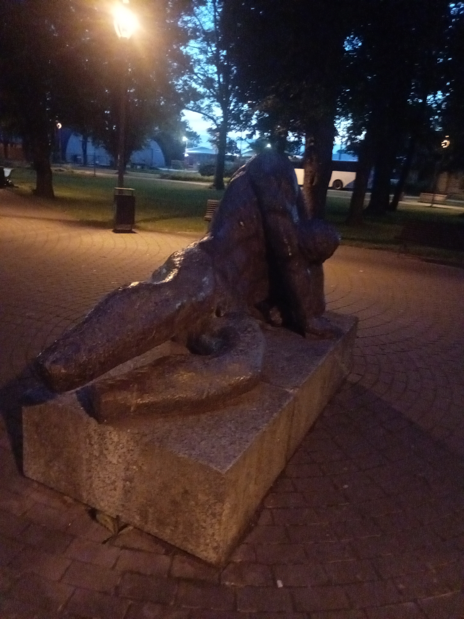 The image shows a bronze sculpture of a nude male figure in a crouching or kneeling position, resting on a large granite base. The sculpture is located in Tapa, Estonia, at an unknown specific location within the city, likely a park or public square, as indicated by the surrounding environment. The figure appears to be in a state of distress or contemplation, with its head bowed and body hunched over. The bronze has a weathered patina, suggesting it has been exposed to the elements for some time.
The surrounding area is paved with brickwork, and there are trees and streetlights visible in the background, suggesting it is nighttime or dusk. The lighting from the streetlights casts long shadows and highlights the texture of the sculpture and the ground. The overall atmosphere is somber and still, with the sculpture being the central focus of the composition. The presence of park benches and a trash receptacle further suggest a public outdoor space. The background is blurred and out of focus, drawing attention to the sculpture.