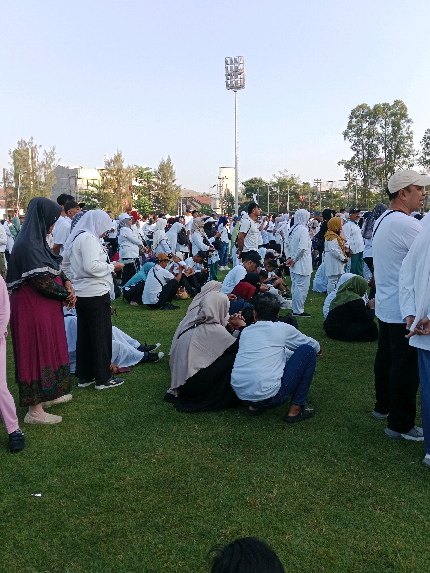 A large group of people, predominantly wearing white attire and headscarves, are gathered on a grassy field in Surakarta, Indonesia. The scene appears to be an outdoor event, possibly a religious gathering or festival, given the clothing and the presence of a large floodlight tower in the background, common in sporting or large public venues. The sky is clear and bright, suggesting daytime. Many individuals are seated on the grass, while others stand or walk. The environment includes trees and some buildings in the distance. The atmosphere is communal, with people interacting and participating in an organized event.
