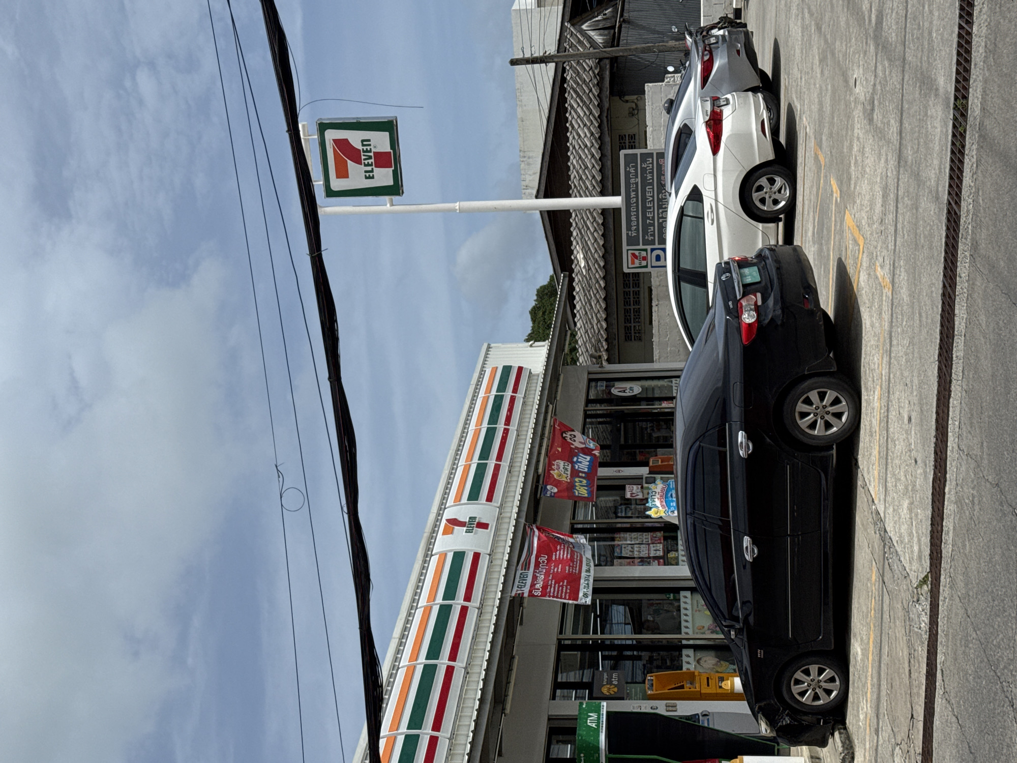 In Rawai, Thailand, a black sedan is parked in front of a 7-Eleven convenience store. The store has a white facade with red, green, and orange striped awnings. A 7-Eleven sign is visible on a pole above the entrance. Several other cars are parked in front of the store, including a white sedan and a silver sedan. The sky is cloudy. There are power lines stretching across the top of the image.