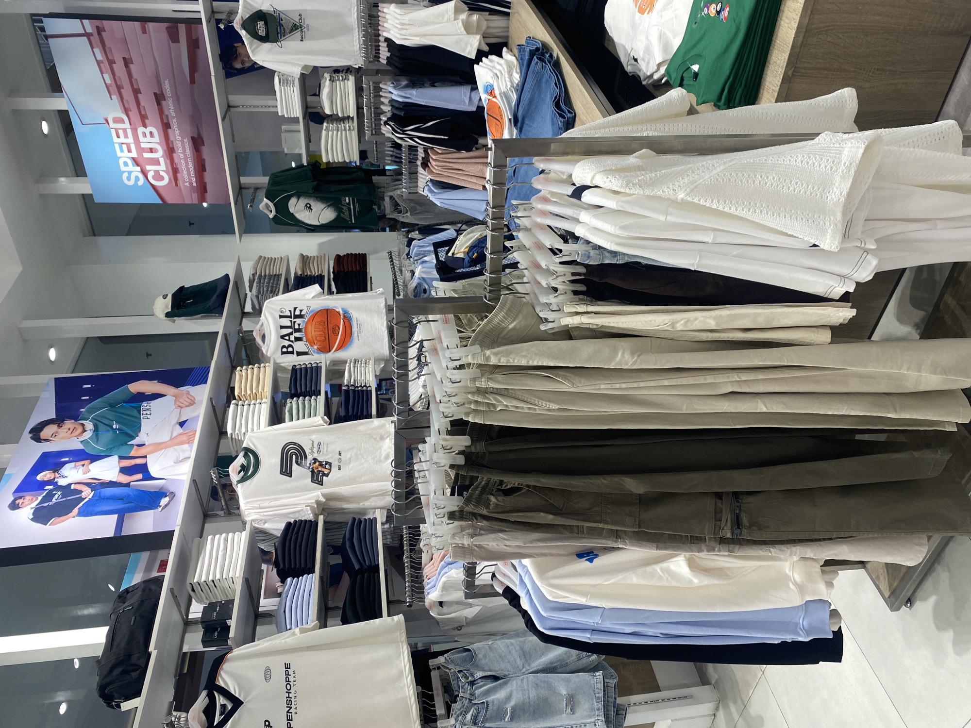 This image captures a brightly lit indoor retail clothing store, likely a Penshoppe branch in Davao City, Philippines. The scene is dominated by racks and shelves displaying a wide array of casual apparel, including t-shirts, hoodies, pants, and jeans.
The clothing predominantly features neutral colors such as white, cream, beige, and black, alongside shades of green, blue, and denim. Many items are neatly folded on shelves, while others hang from white plastic hangers on metal racks. Notable garments include white t-shirts with graphics like a basketball and the text "BALL LIFE CLUB," and green t-shirts with a rugby ball design. A section of white, textured long-sleeved tops is also visible.
Large promotional posters are affixed to the walls on the left. One poster features models wearing Penshoppe apparel, with text like "PENSHOPPE" and "Lightspeed." Another prominent blue and red sign advertises "SPEED CLUB," described as "A collection of bold graphics, athletic codes. and modern classics." Additional visible text on clothing includes "PENSHOPPE RACING TEAM" and "R-Club." The store appears modern and well-organized, with overhead lighting illuminating the merchandise.