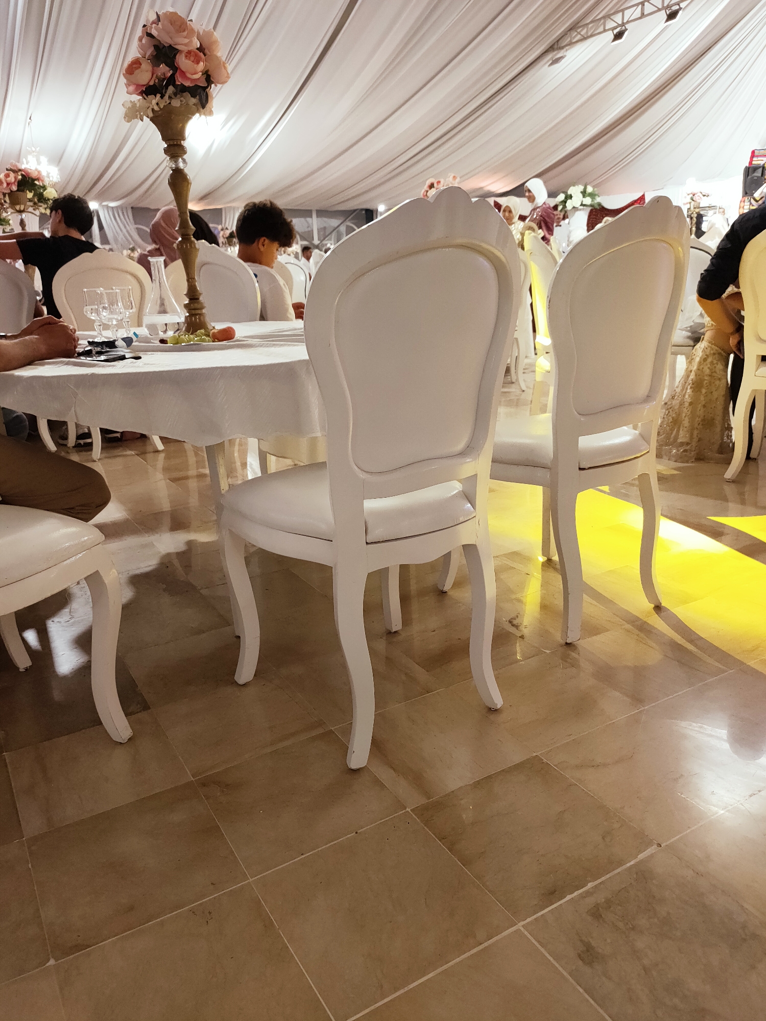 The image depicts an indoor event space, likely a banquet hall, with white draped ceilings and tiled floors. Several tables are set with white tablecloths, chairs, and floral centerpieces. People are seated at the tables, engaged in various activities such as eating and socializing. The scene is illuminated by warm lighting, creating a festive atmosphere. The location is identified as Mornag, Tunisia, suggesting a wedding or similar celebration is taking place in this North African setting.