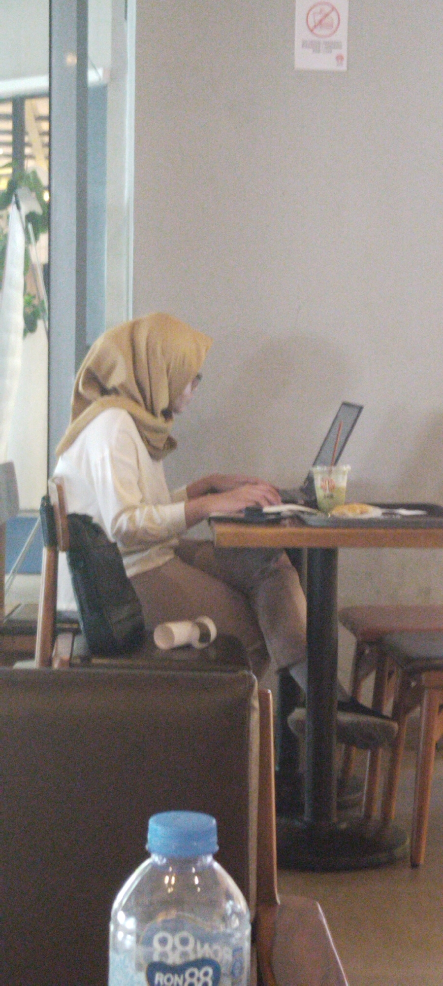 A woman wearing a tan hijab and a white long-sleeved shirt sits at a table, typing on a laptop. A green beverage in a clear cup with a straw is on the table, along with a plate containing a pastry. A white tube-shaped object rests on the seat of the chair beside her. The scene appears to be indoors, possibly a cafe or restaurant. In the foreground, a partially visible clear plastic water bottle with a blue cap and a "RON88" label is present. A "No Smoking" sign is visible on the wall in the background. The overall lighting is dim, suggesting an indoor setting.