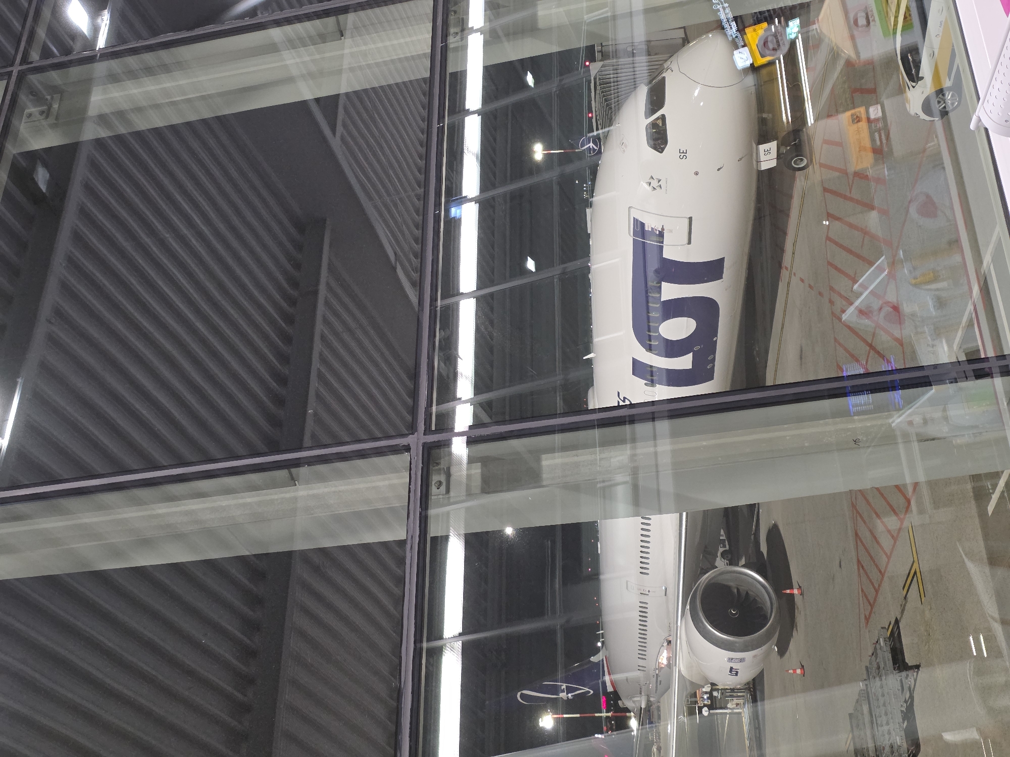 An aerial view shows a parked LOT Polish Airlines airplane at an airport terminal at night. The airplane is white with large blue LOT lettering across the fuselage and a blue and white logo on the tail. The plane is connected to a jet bridge and is surrounded by various airport equipment and vehicles, including a baggage cart and a small white airport service vehicle. The airport terminal has a large glass facade through which the airplane is visible, and the ceiling of the terminal is ribbed and gray. The scene is lit by artificial lights, reflecting off the glass.
