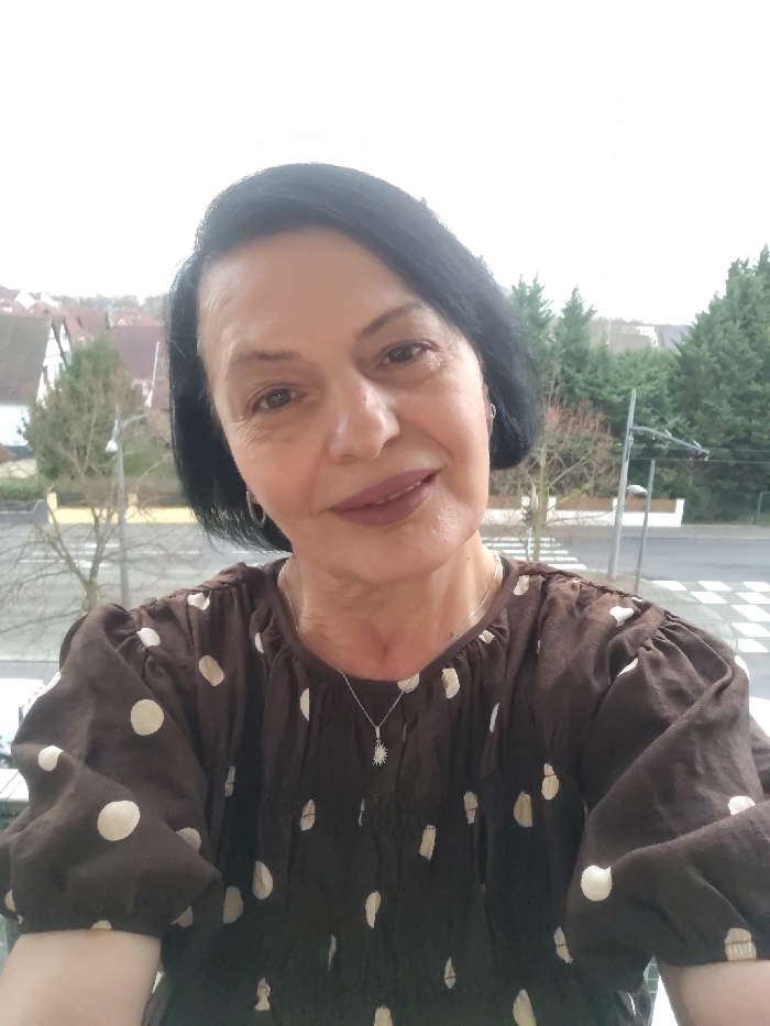 A woman is centered in the frame, wearing a brown, long-sleeved polka-dot shirt and a silver pendant necklace. She has dark, short hair and is wearing lipstick. Outside, a cityscape is visible in the background. There are trees and buildings on the horizon. A street with a crosswalk is directly behind the woman. The sky is overcast.