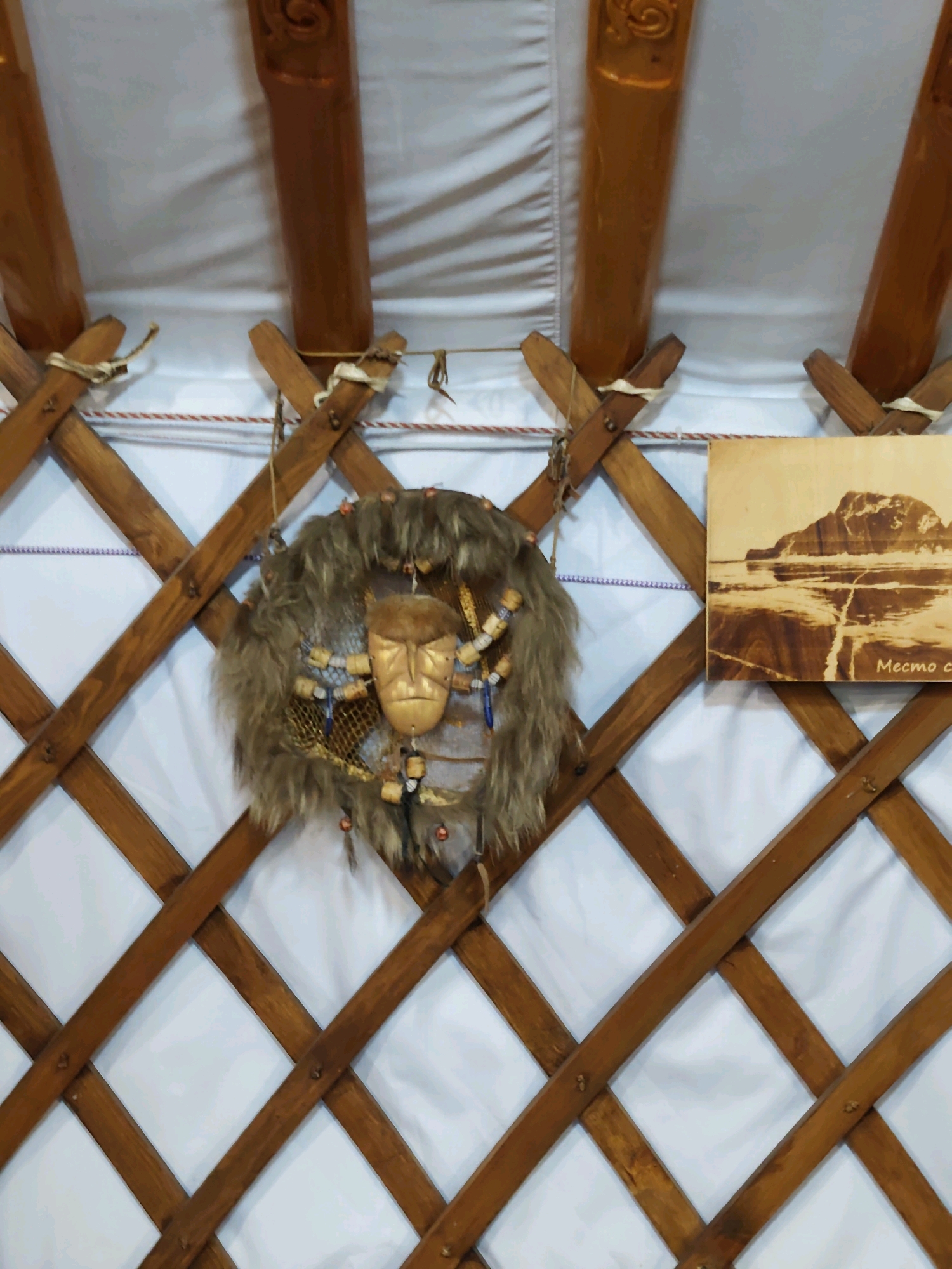 This is a close-up, eye-level, indoor shot of the interior of a traditional yurt in Listvyanka, Russia. The yurt has a latticework wooden frame, visible on the walls, and wooden poles extending towards the ceiling. The white fabric lining of the yurt is also visible.
Central to the image is a decorative mask hanging on the latticework. The mask is framed by a circular arrangement of brown fur, beads, and small decorative elements. The mask itself appears to be made of wood or a similar material, with carved facial features and a golden hue.
To the right of the mask, a framed image is attached to the latticework. The image is a sepia-toned photograph or print depicting a rocky landscape with water or ice in the foreground. The text "Место с" is visible in the bottom right corner of this framed image.