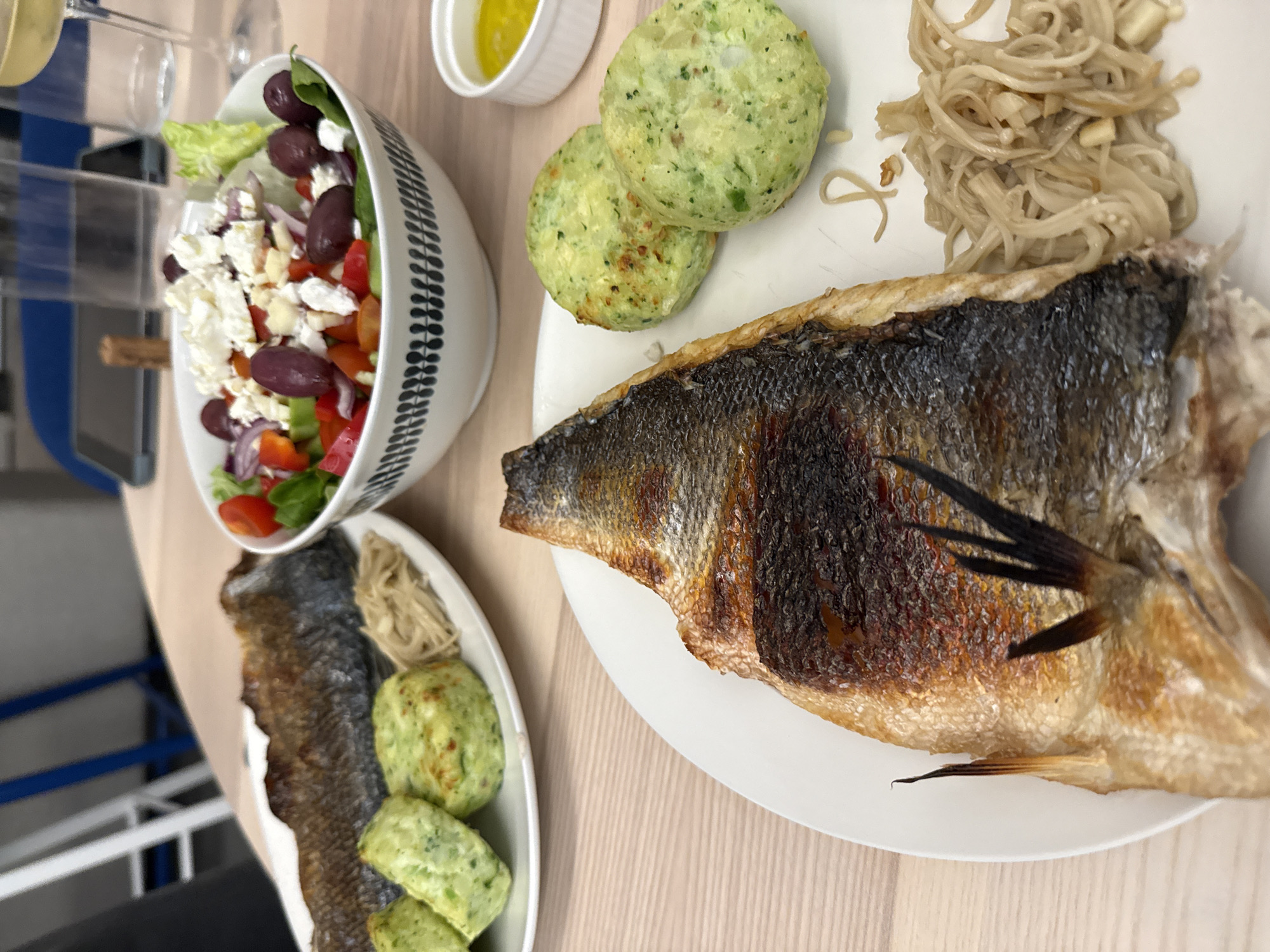 The image shows a meal served on a wooden surface, possibly in Livadia, Larnakas, Cyprus. A Greek salad is in a white bowl with a leaf-patterned rim, containing lettuce, tomatoes, red onions, olives, and feta cheese. A small white dish contains a yellowish liquid, likely olive oil or a vinaigrette. On a white plate, there is a whole grilled fish with crispy skin, along with two green patties, and a small pile of noodles. A similar meal is also served on a small plate. The plates are located indoors. A few glasses can be seen. The objects are oriented as if on a table ready to be consumed.