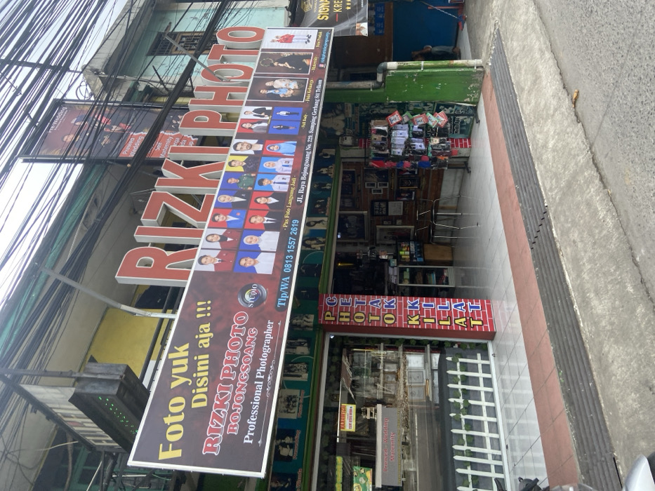 The image shows the storefront of a photography studio named Rizki Photo, located at JL. Raya Bojongsoang No. 223, Sampling, Gerbang Sit. Tekan, Bojongsoang, Indonesia, according to the sign. The red, yellow, and white sign is partially obscured by wires and reads "Foto yuk Disini aja!! Rizki Photo Bojongsoang Professional Photographer Studio Tlp/WA 0813 1557 2619." There are many framed photographs of people, some in formal attire, displayed in the storefront windows. Several chairs are visible inside the studio. The sidewalk in front of the studio is paved with concrete and has a ramp leading to the street. A motorcycle is parked on the sidewalk in front of the store. The storefront is decorated with green painted brick and green plants. The building's exterior is grey. The studio is located on a busy street, with many other buildings and businesses nearby. Wires and electrical lines are strung across the street. The image was likely taken on a sunny day.