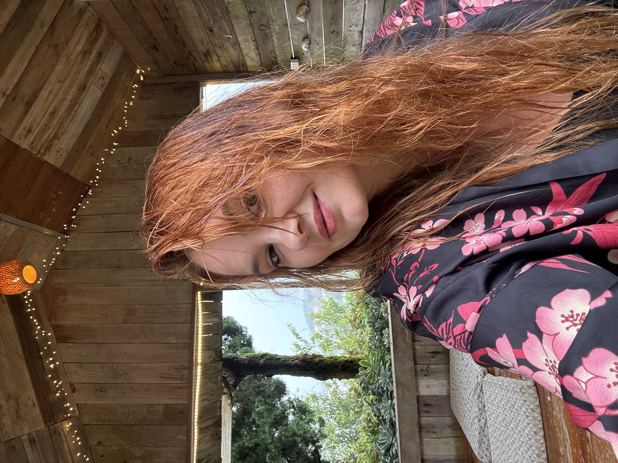 A woman with long, red, wet hair is taking a selfie indoors. She is wearing a dark robe with a pink floral pattern. Her hair is partially covering her face. The background is a wooden structure with a sloped roof adorned with string lights and a hanging lantern. Through an opening, a misty, green landscape with trees and foliage is visible. The overall atmosphere appears to be cozy and perhaps a bit rustic. The location is the city of Lộc Quý, Vietnam.