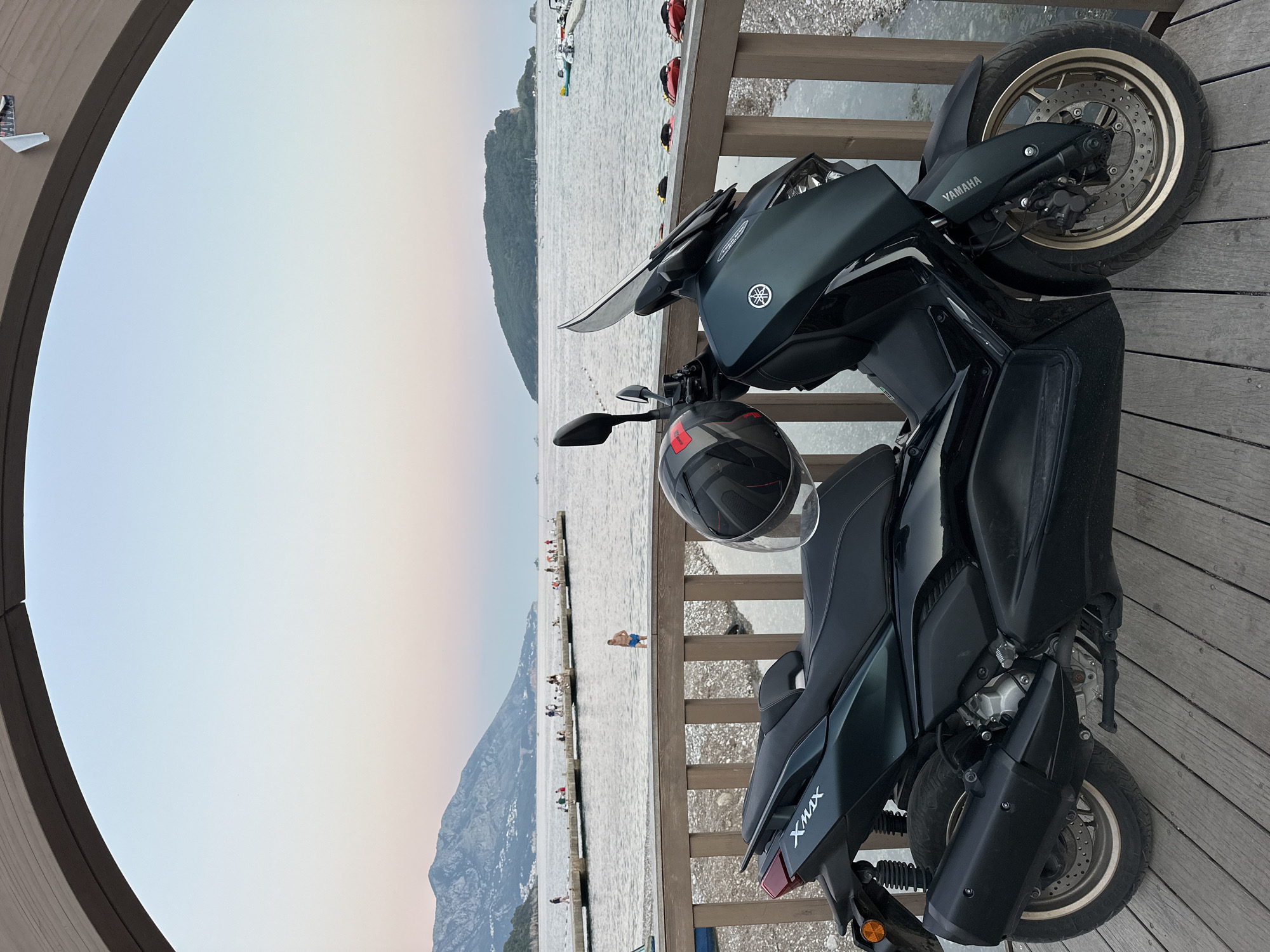 The image depicts a dark green Yamaha XMAX scooter parked on a wooden deck overlooking a scenic coastal view at dusk. The scooter is positioned in the foreground, with its front wheel slightly turned. A black helmet with red accents rests on the scooter's seat.
In the background, a long pier extends into the calm sea, where several people are visible fishing or relaxing. Further out, mountains line the horizon, their peaks softly illuminated by the twilight sky. An island, covered in lush greenery, is situated in the middle distance. The sky transitions from a pale blue at the top to a soft gradient of pink and orange near the horizon.
The location is identified as Budva, Montenegro. The overall atmosphere is serene and peaceful, suggesting the end of a day at the coast.