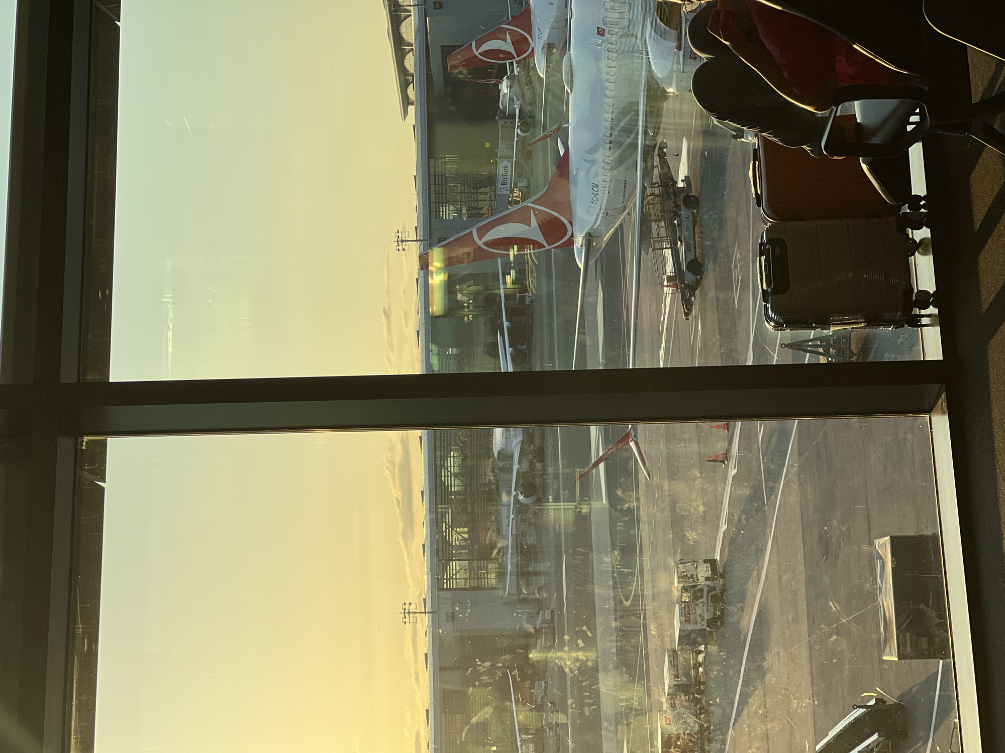 The image depicts an airport scene during sunset, viewed from inside a terminal through a large window. Several airplanes are visible on the tarmac, some parked at gates and others further away. One airplane in the foreground prominently displays the Turkish Airlines logo on its tail fin. Various airport service vehicles, including baggage carts and tugs, are positioned on the tarmac.
In the lower right portion of the image, passengers are seated in an airport waiting area. Luggage, including a dark gray rolling suitcase and an orange suitcase, is placed near the seats. The setting sun casts a warm, golden light across the scene, illuminating the sky with hues of yellow and orange. The architecture of the airport terminal is visible in the background, with glass facades reflecting the sky and surrounding aircraft.
The location is identified as Istanbul Airport, which is situated in the Arnavutköy district of Istanbul, Turkey. The overall atmosphere is one of travel and anticipation, characteristic of an airport environment at the end of the day.