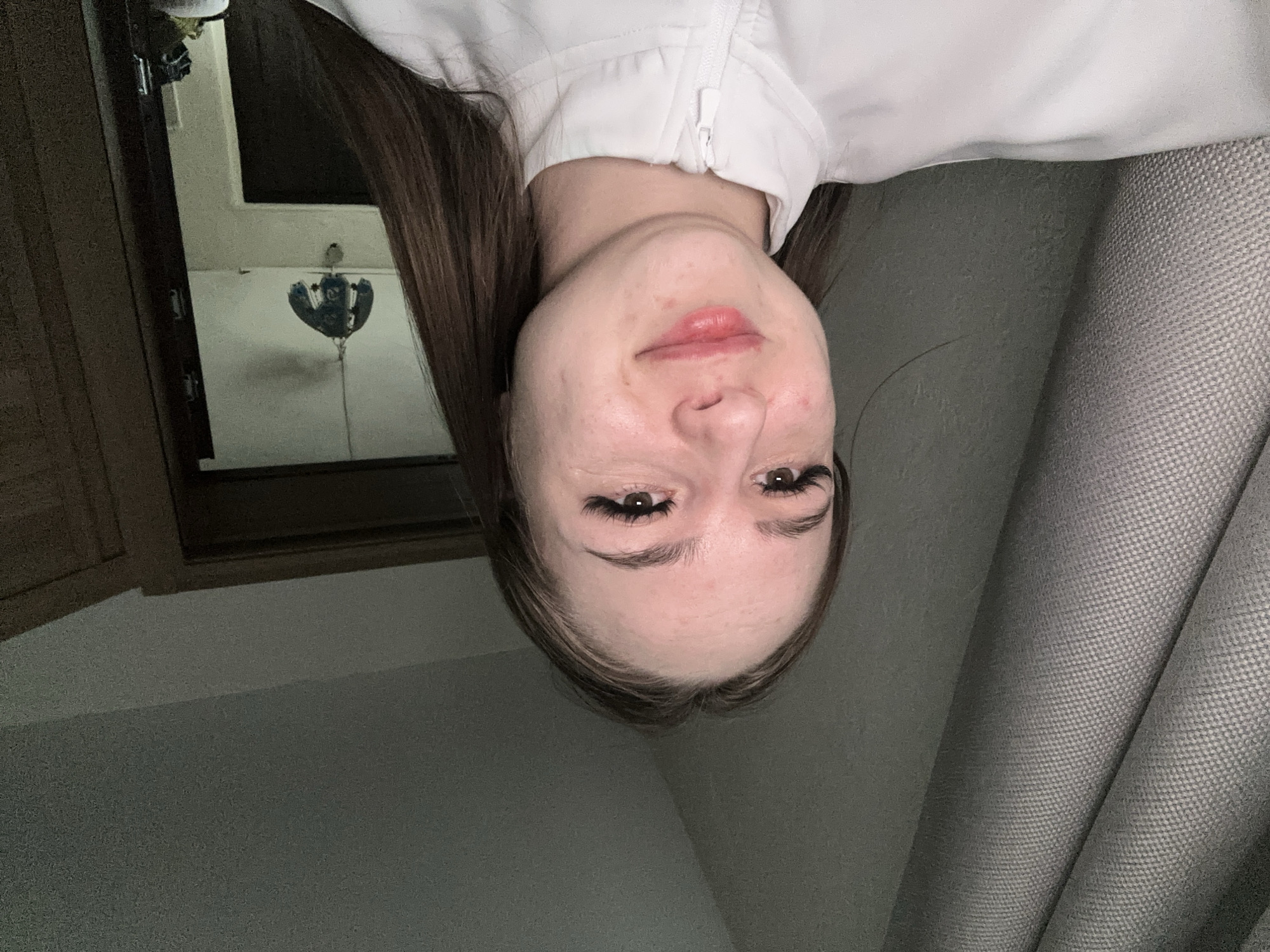 A selfie taken by a Caucasian female, upside down. She is wearing a white zippered shirt. Behind her on the left is a dark wooden door frame and window. In the window is a small mobile. To her right is a gray curtain. The location is Unknown, Unknown.