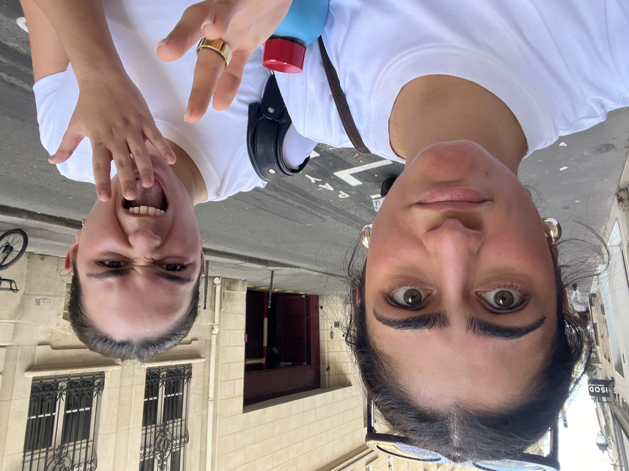 The image shows two individuals in Paris, France. They are facing the camera, which is held upside down. Both are wearing white t-shirts. The individual on the left is expressing a playful, exaggerated expression, while the individual on the right maintains a more neutral expression.
The location shows a Parisian street. Architectural details, including buildings with windows with iron railings, are visible in the background. Part of the sidewalk and street are also visible. A bicycle is leaning to the left in the corner of the image.