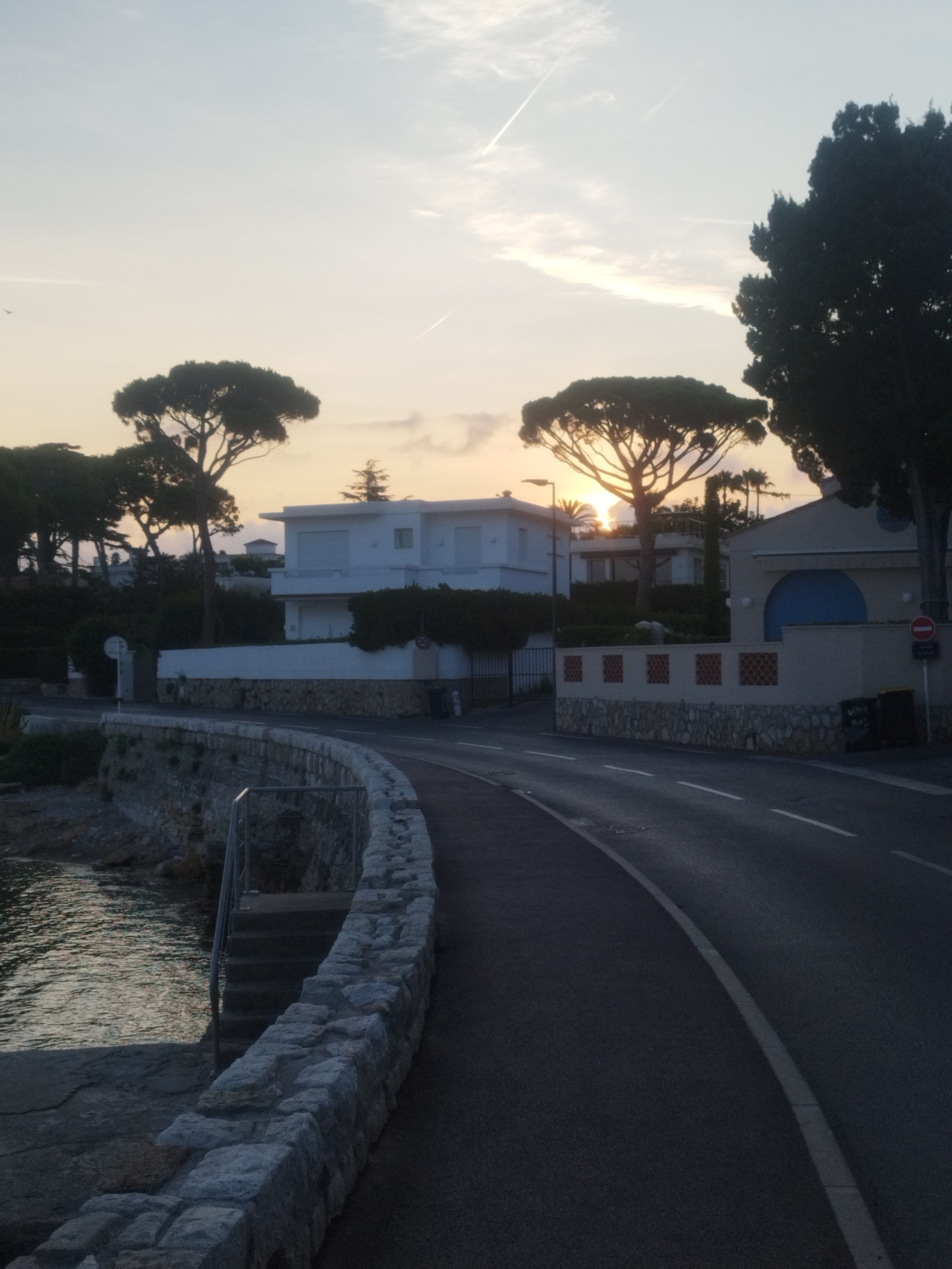 The image depicts a scenic coastal road in Antibes, France, at dusk. The sun is setting, casting a warm glow across the sky and illuminating the tops of pine trees that line the road. The road curves to the right, with a stone wall bordering the sea on the left. The sea itself appears calm, with gentle ripples reflecting the sky.
Several houses, typical of the French Riviera, are visible in the background. They are predominantly white with modern architectural styles, featuring balconies and large windows. Some houses are partially obscured by the lush green foliage and tall trees.
On the roadside, there are a few objects: a street lamp, a trash bin, and a circular traffic sign indicating a speed limit of 30 km/h. The road is asphalt and appears to be in good condition, with white lane markings visible.
The overall atmosphere is tranquil and serene, suggesting a peaceful evening by the Mediterranean Sea. There are no people visible in the image, enhancing the sense of quiet solitude. The sky is a soft gradient of blue and orange, with a few wispy clouds and contrails from airplanes.