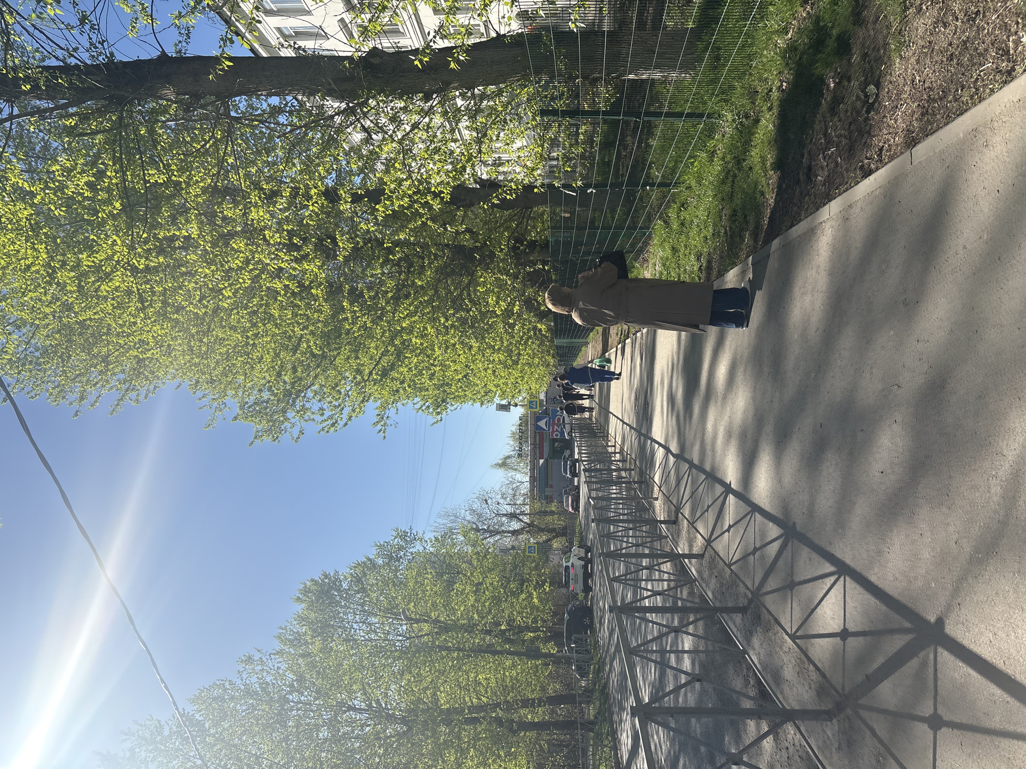 The image depicts a pedestrian walkway in Perm, Russia, on a sunny day. The walkway, lined with trees bearing fresh green leaves, runs alongside a metal fence separating it from a grassy area. A woman in a tan coat walks on the path, her back facing the camera. Other pedestrians are seen further down the walkway. Cars are parked on the street parallel to the walkway. The sky is clear with a visible sun glare. Building facades can be seen in the background.