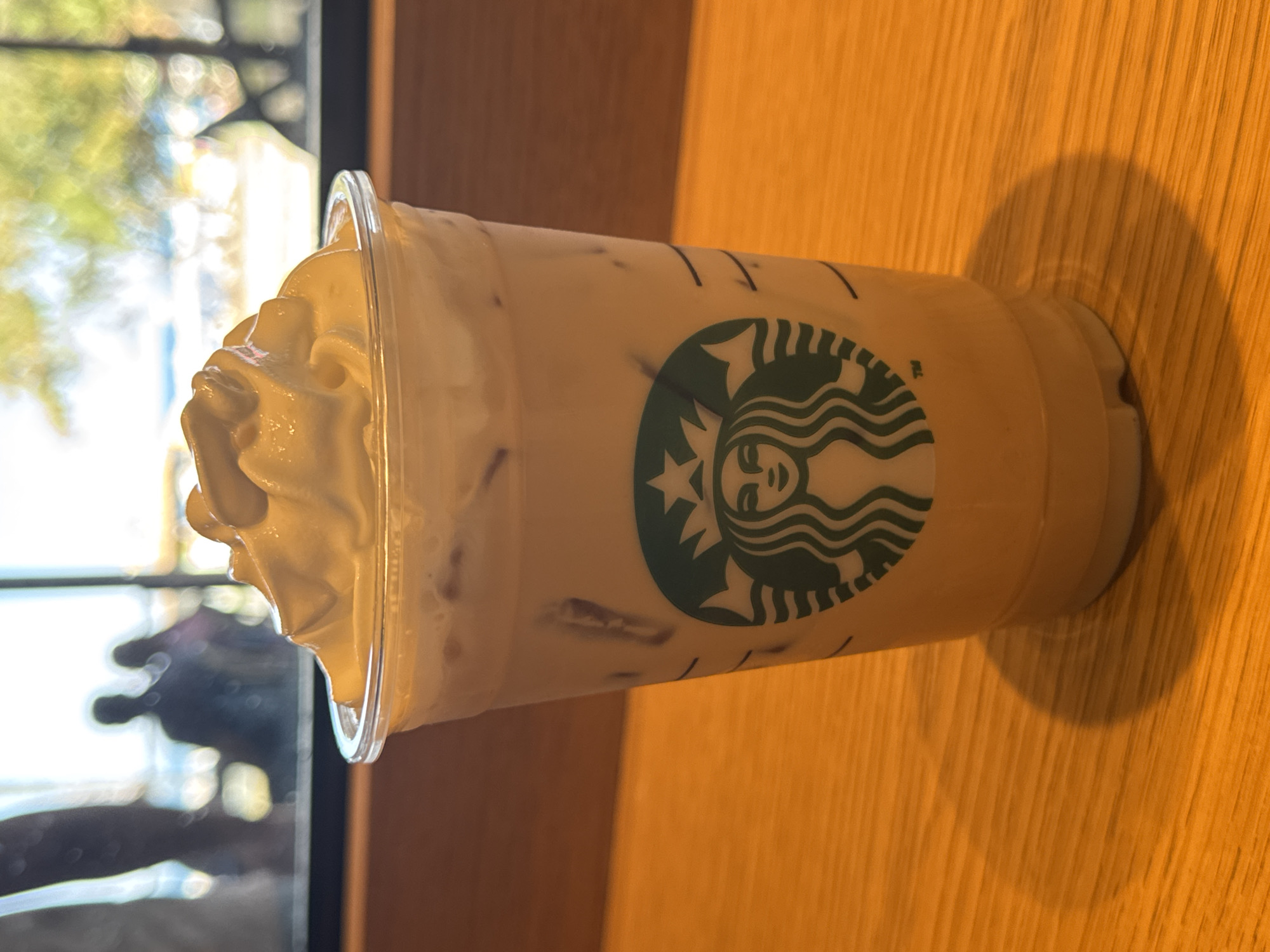 An iced coffee drink with whipped cream is in a clear plastic cup with the Starbucks logo. The drink is on a wooden table. In the background, out of focus, are silhouettes of people visible through a window, with trees and sky beyond. This scene is not identifiable as being in Kemer, Turkey.
