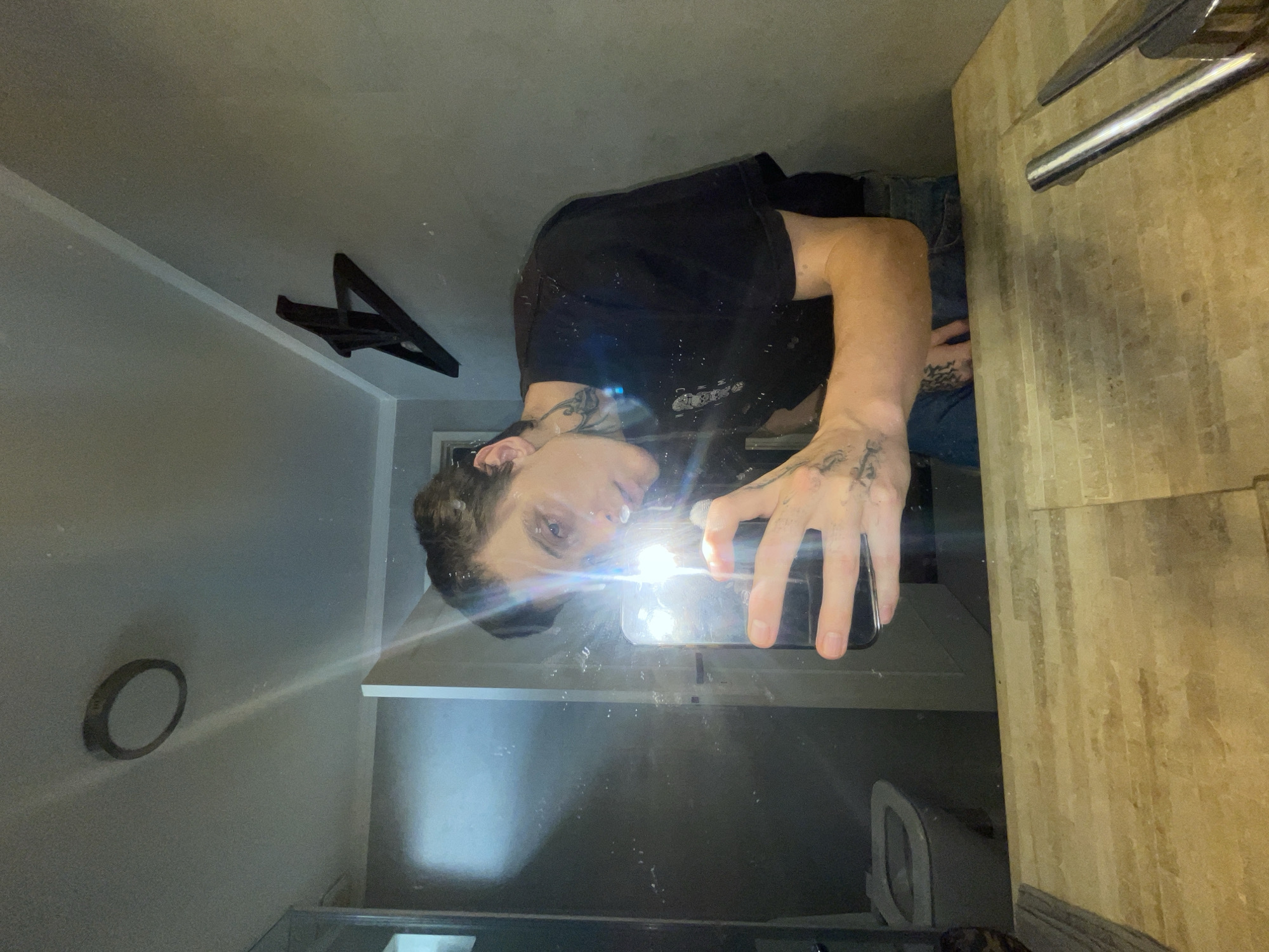 A person is taking a mirrored selfie in what appears to be a bathroom. The person is wearing a black t-shirt and jeans, and has tattoos on their arms and hands. The bathroom has a white sink and cabinet, with a wood-paneled wall to the right. In the reflection, a toilet is visible. There is a round object on the wall, and a black triangular decor on the ceiling. The location in the city of Vicente López, Argentina cannot be determined from the image.