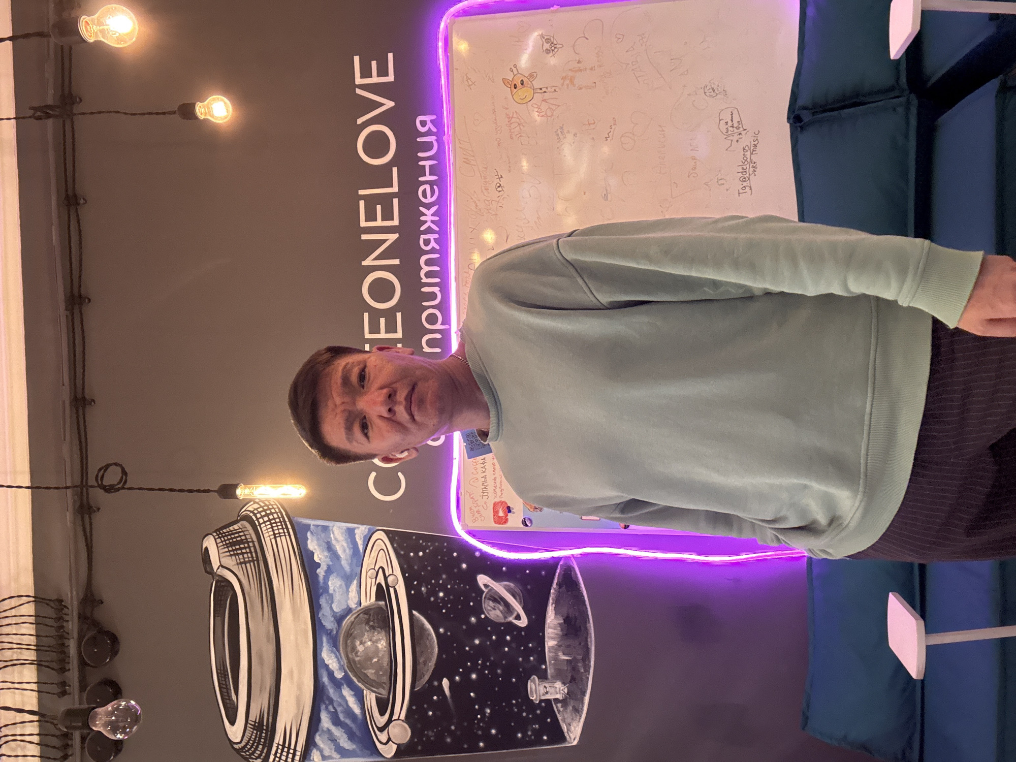 A medium shot features a man in front of a branded wall. He is wearing a light blue sweatshirt and striped pants. The wall displays "ONE LOVE" in large white letters and "притяжения" in a smaller, vertical format. The backdrop is illuminated by a purple neon light. Below, a graphic of planets and space decorates the wall. Above, a whiteboard is covered with signatures and drawings. Decorative light bulbs hang from the ceiling above. The location is likely Moscow, Russia, given the Cyrillic text.