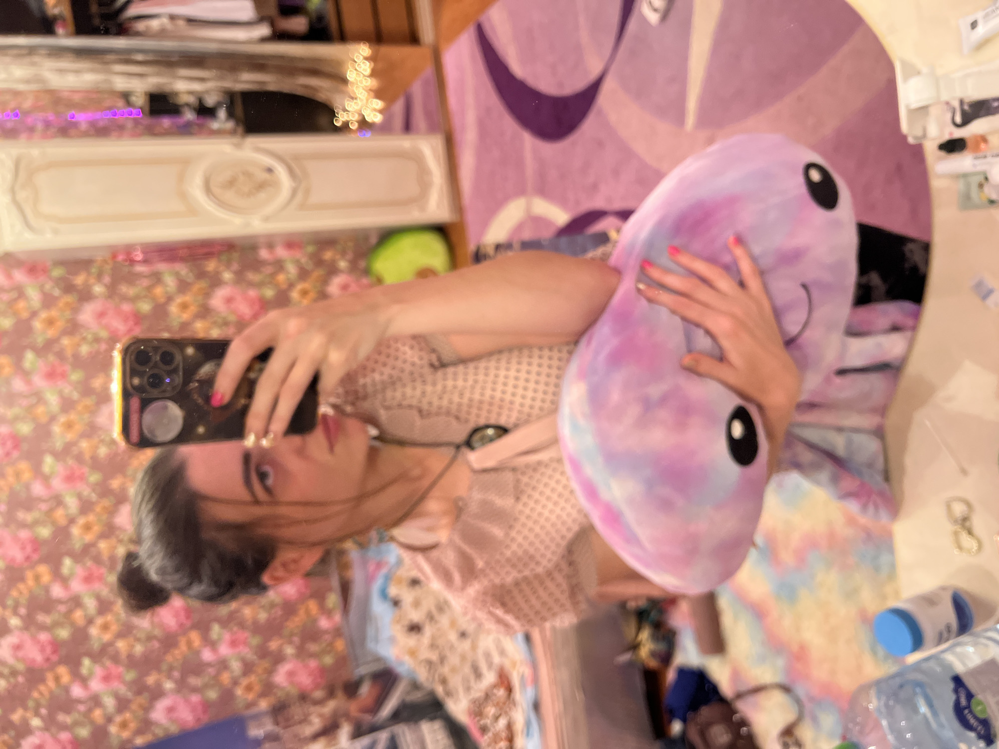 The media file shows a reflection in a mirror of a young person holding a stuffed toy. The person, with dark hair pulled back, is wearing a light-colored patterned top. They are holding a large, purple and pink tie-dyed stuffed toy that resembles an alien or an octopus with large black eyes. The person is taking a selfie with a smartphone that has a gold case.
The background reflected in the mirror is a bedroom or dressing area. To the left, there is a floral wallpaper with pink and orange flowers. Above the wallpaper, a white ornate shelf or headboard is visible. In the center of the reflection, a large purple and white patterned tapestry or fabric hangs on the wall, creating a whimsical backdrop. Scattered on a surface to the right are various items including what appears to be toiletries, a key ring, and a bottle of water. The lighting suggests it might be daytime, as there is ample light, but the exact time is not discernible. The overall mood is casual and playful, possibly indicating a moment of relaxation or self-expression. No visible text is clearly legible within the frame. The location context of Krasnoe-on-Volga, Russia, is provided but cannot be visually confirmed from the image itself.