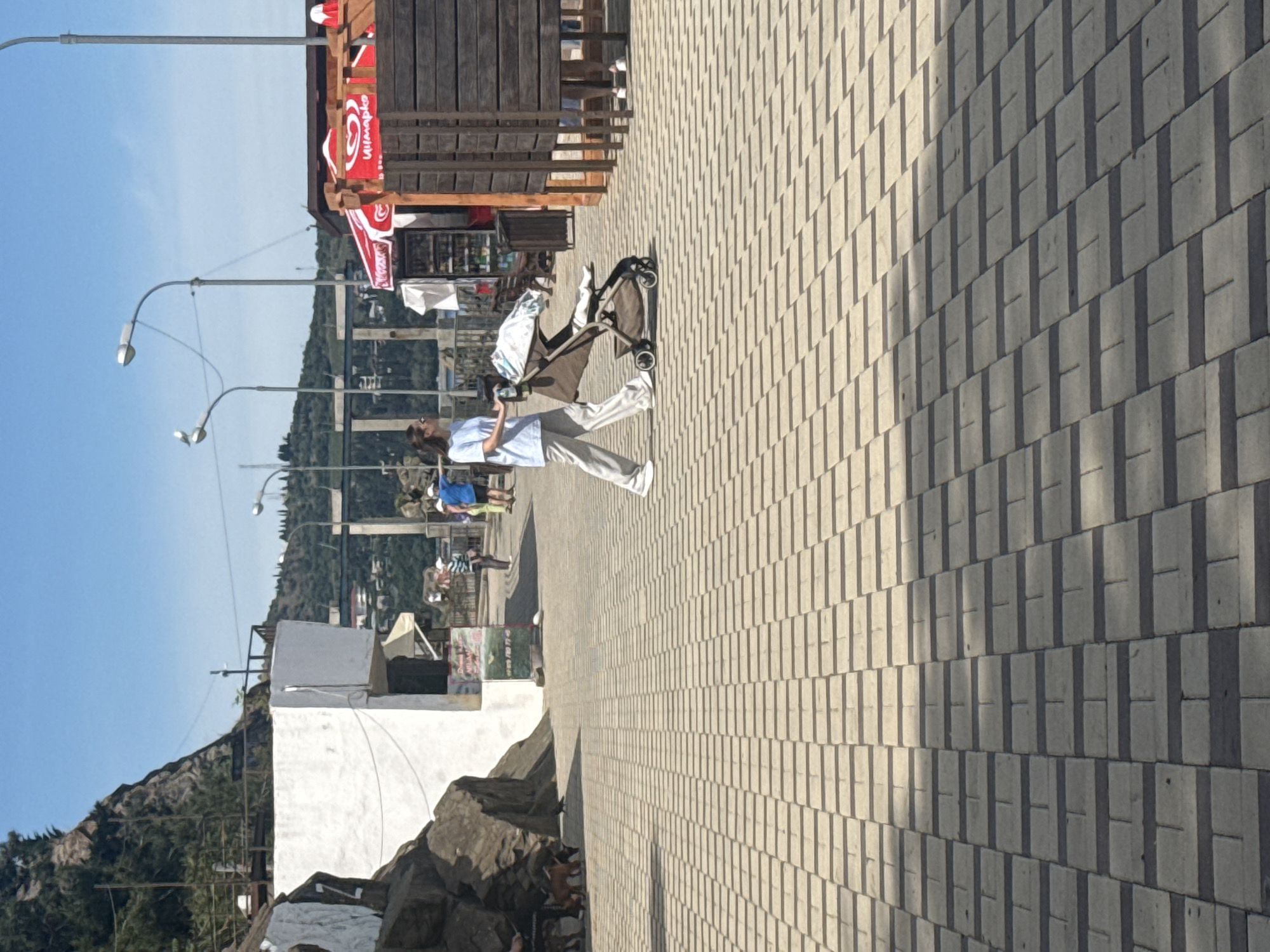 The image depicts an outdoor pedestrian area on a sunny day, oriented as a paved walkway leading into the distance. The ground is covered with light-colored rectangular paving stones, exhibiting strong shadows from direct sunlight.
A woman is prominently featured on the right side of the path, walking and pushing a dark-colored baby stroller with a light-colored covering. She has dark hair and is dressed in a light blue long-sleeved shirt, light-colored pants, and white sneakers.
To the left of the path, a series of wooden structures, appearing to be shops or stalls, line the walkway. Several red banners are attached to these structures, displaying a white heart logo with the brand name "Cornetto" clearly visible, indicating an ice cream vendor. Further in the background, indistinct figures are visible near these stalls.
The background features a clear blue sky above a landscape of green, forested hills. In the foreground on the far left, there is a white building next to a rocky area, with more hills behind it. Several lampposts are spaced along the pathway.
The strong, defined shadows cast by the lampposts, the woman, and the stroller suggest it is midday or early afternoon under bright, clear weather conditions. The overall scene conveys a leisurely atmosphere in a public, possibly commercial or tourist-oriented, outdoor setting.