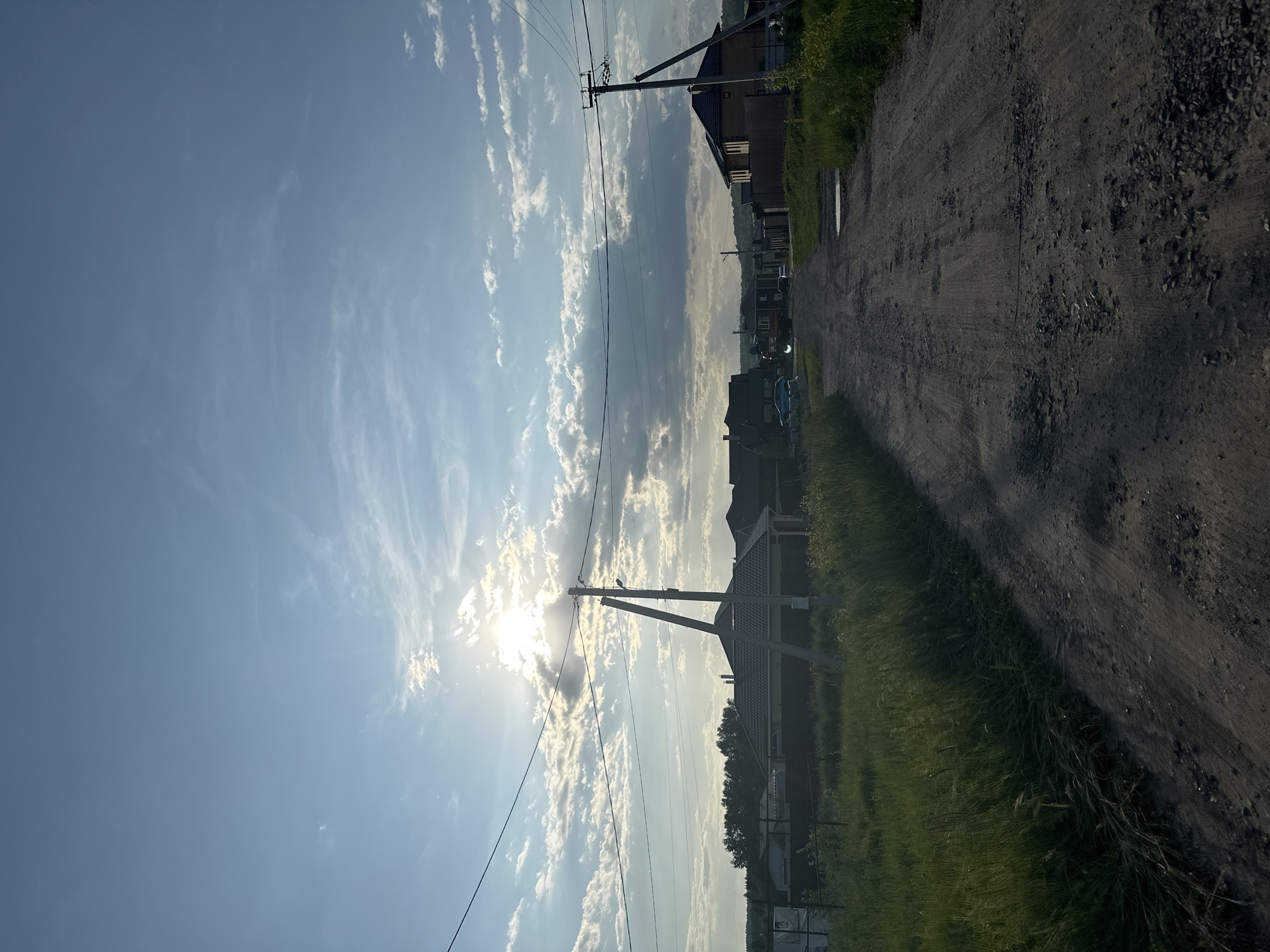 The image depicts a rural dirt road leading into a residential area under a partly cloudy sky. The sun is partially obscured by clouds, casting rays of light. Power lines stretch across the scene, supported by utility poles. On either side of the road, there is tall green grass and some vegetation. Several houses with visible roofs are situated along the road. In the background, a treeline is visible, and a slight incline suggests a gentle hill. There are no people or animals visible in the frame. The overall impression is that of a quiet, sunlit day in a village setting. The location is the village of Estachi, Russia.