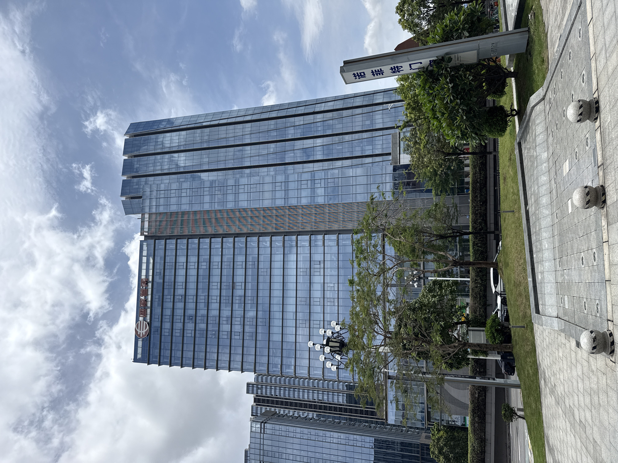 The image shows a modern glass skyscraper in Tianhe District, China, under a cloudy sky. The building features a distinctive stepped top and vertical glass panels. A sign with Chinese characters is visible on the right. In the foreground, a paved area with decorative bollards leads to a grassy area with trees and streetlights. Several cars are parked on a road in the background, and other glass buildings are visible to the left. The scene appears to be a commercial or office district in a city.