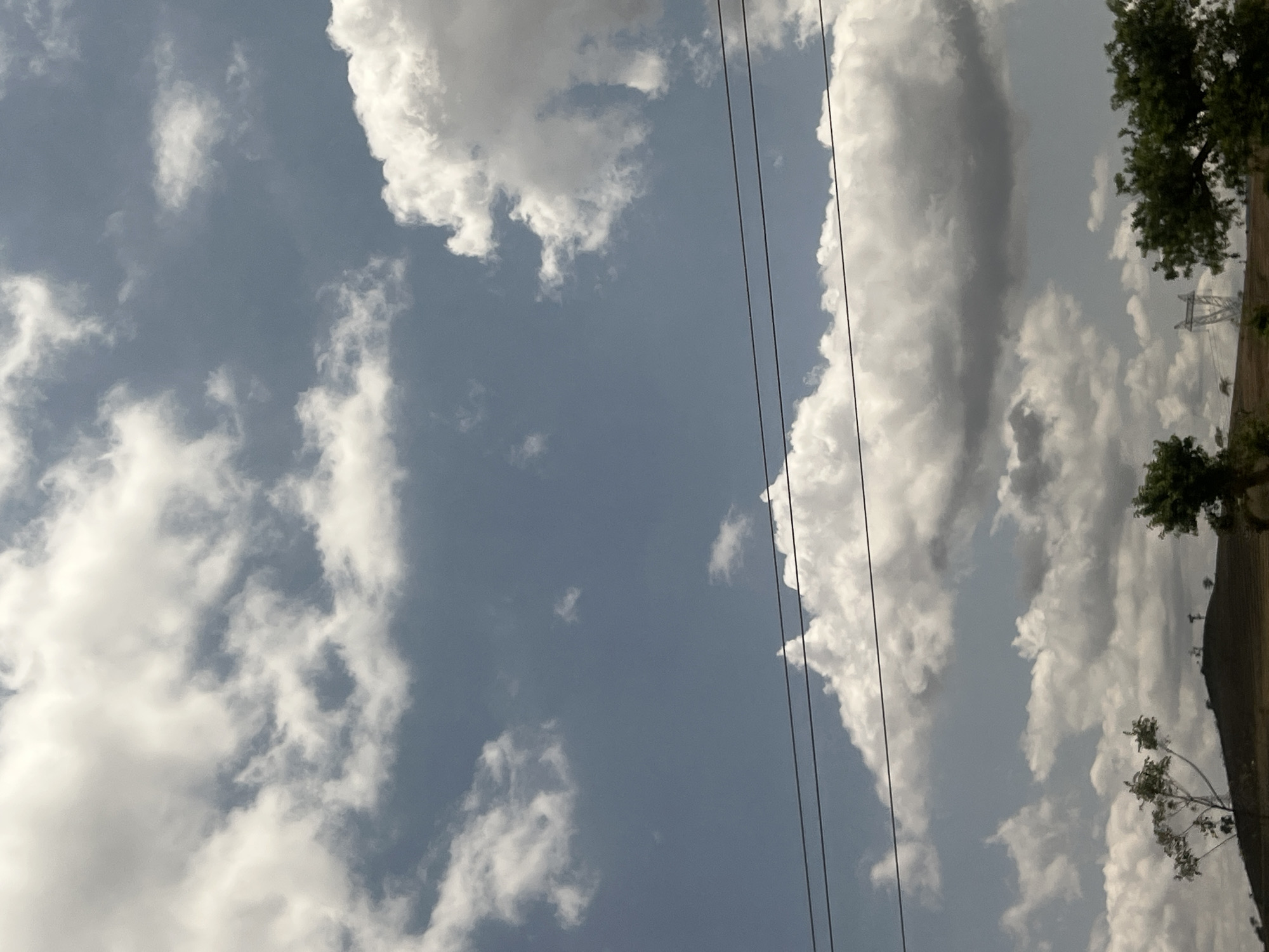 The image captures an outdoor daytime scene in Sabon Layi Jessu, Nigeria, characterized by a partially cloudy sky and a rural landscape. The upper two-thirds of the frame consist of a light blue sky populated by numerous cumulus clouds, ranging in color from bright white, indicative of direct sunlight, to darker gray. Three prominent black utility power lines stretch horizontally across the middle of the image. Below the power lines, the terrain features a large, brown, arid-looking hill or mound in the mid-ground, exhibiting sparse vegetation. In the foreground, on the left, are trees with lighter-colored trunks and green foliage, while on the right, denser green tree canopies are visible. On the horizon, a metal lattice transmission tower is identifiable on the far right, partially obscured by foreground trees, with smaller, less defined utility structures present on the hill to the left. No human or animal subjects are present in the scene.